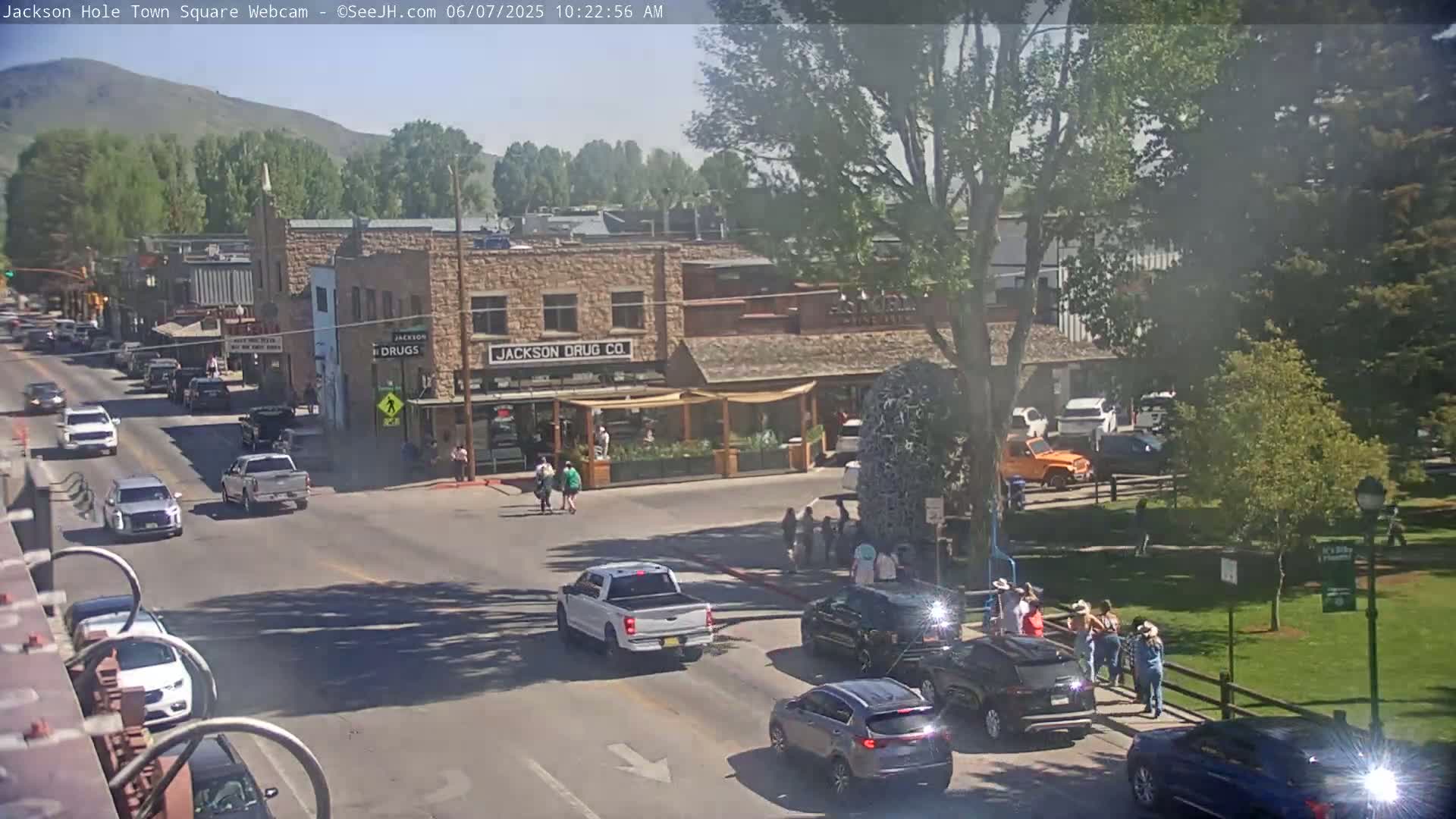 A sunny day reveals a town square with cars driving on roads, pedestrians walking on sidewalks, and buildings lining the streets, including a drug store.