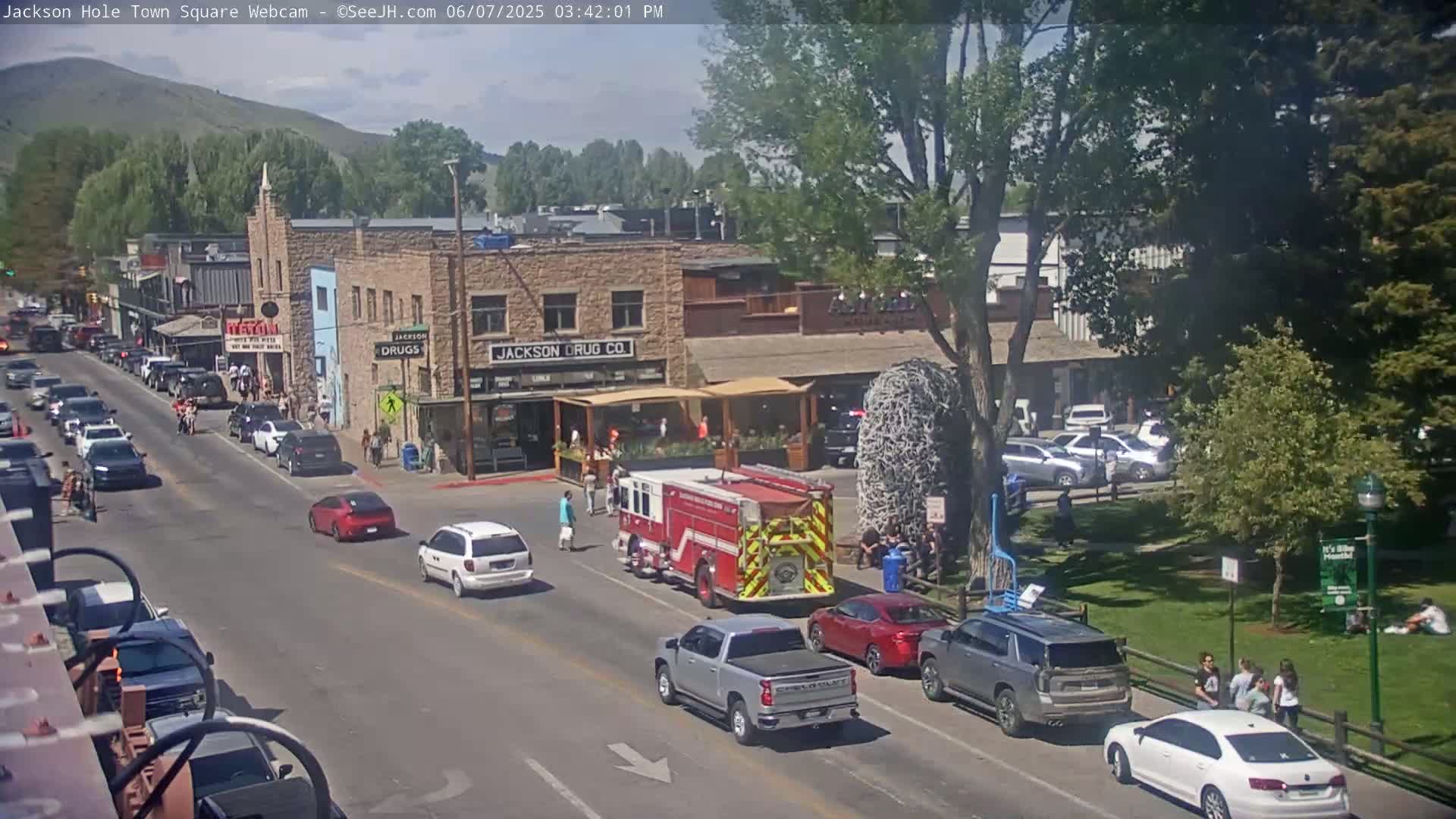 A sunny day in a town square shows a firetruck parked on a street among several cars, with pedestrians and buildings lining the street and a grassy area with people sitting on the grass.