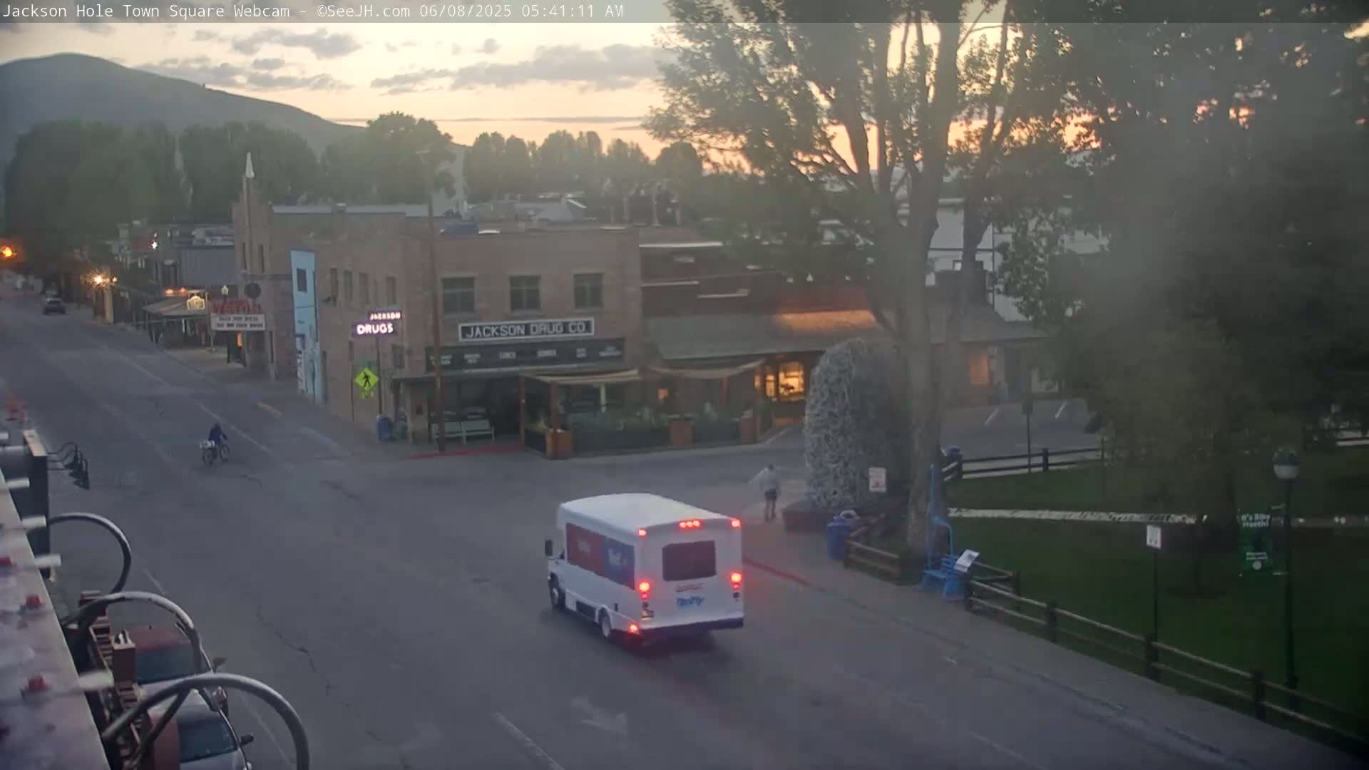 A white shuttle bus drives down a street in a town at dawn, with a cyclist and a person walking nearby, under a hazy sky.