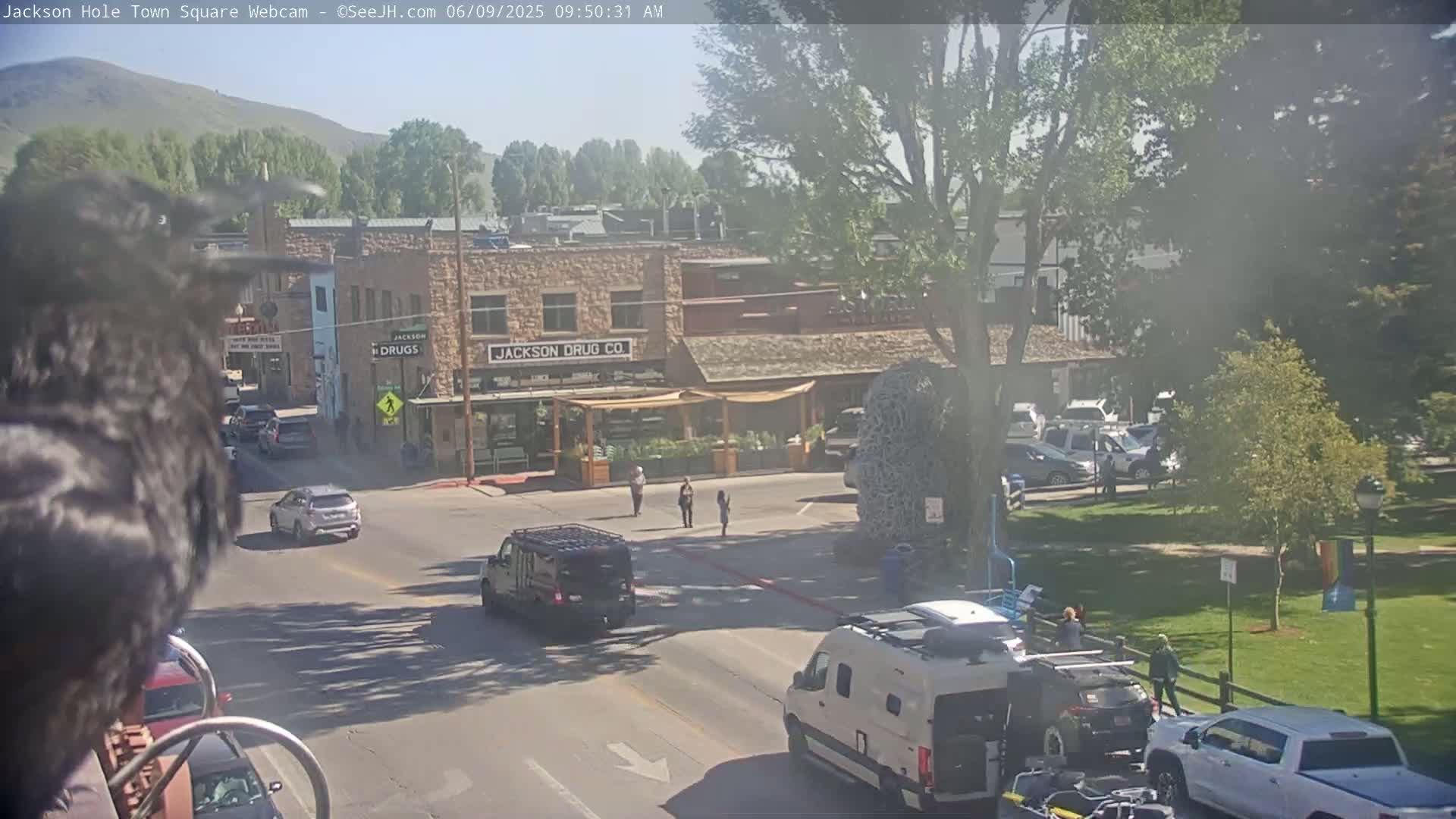 A sunny town square scene shows several vehicles at an intersection in front of stone buildings, with people walking on the sidewalk and a large woven sculpture in the middle of the square.
