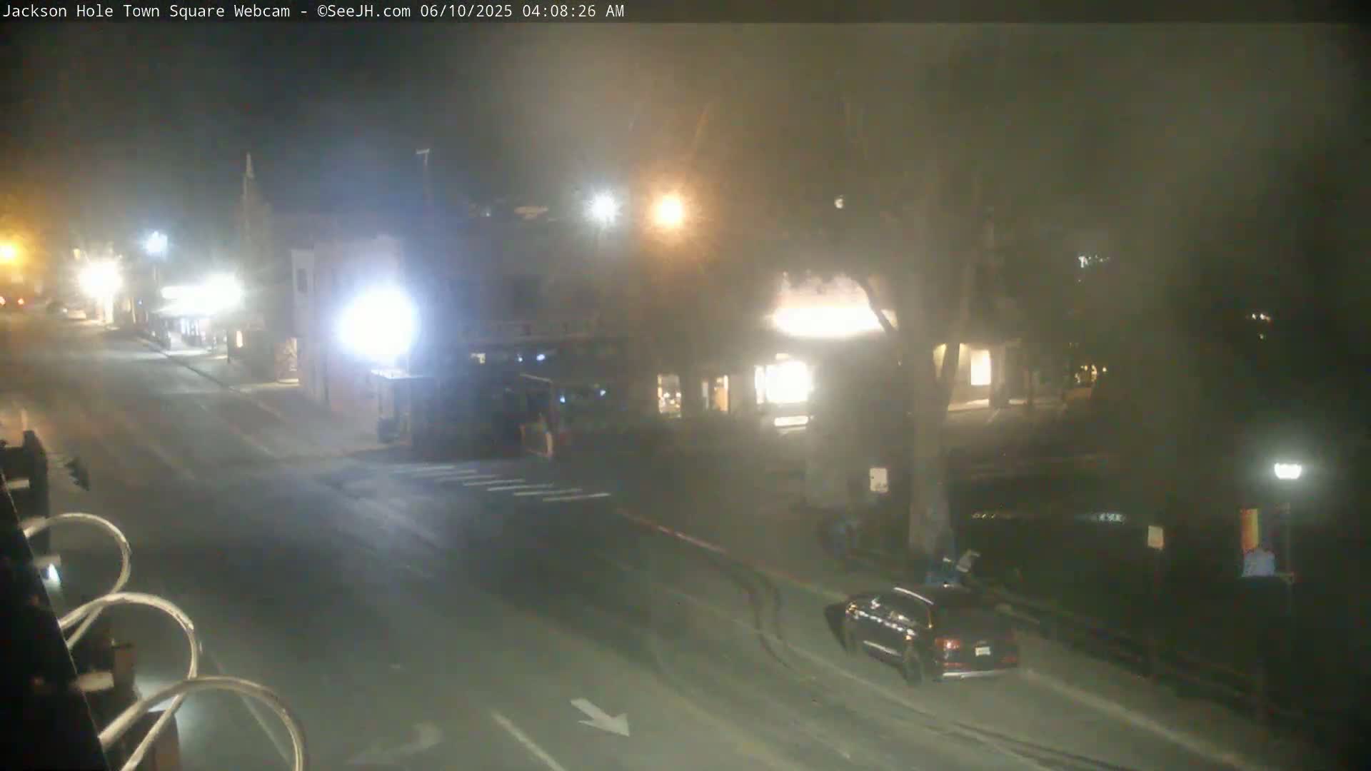 A nighttime, slightly hazy view of a mostly empty street with a few cars and pedestrians near buildings.