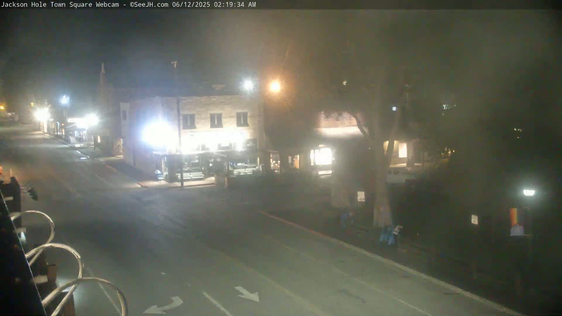 A nighttime, hazy view of a mostly empty street in front of several buildings, with a few people visible on the sidewalk.