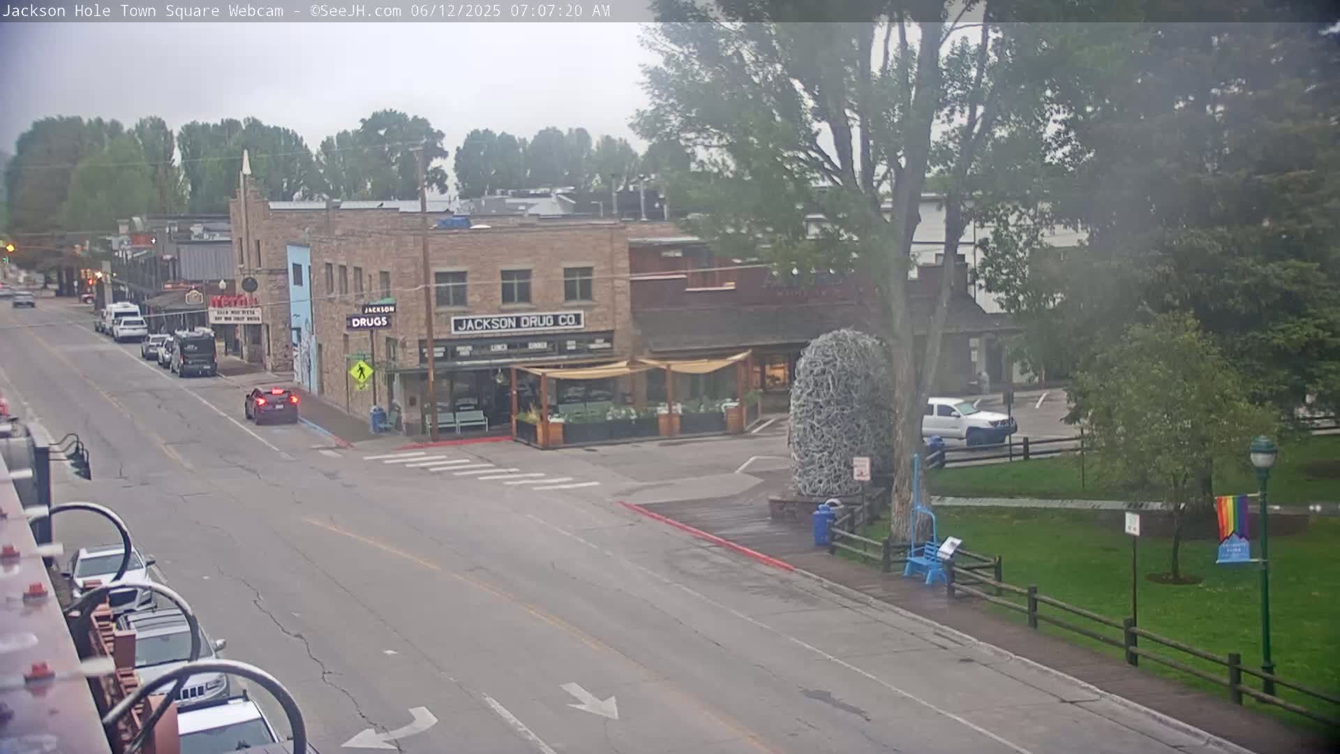 A mostly overcast view of a town square, with cars parked along the street and a few vehicles driving, shows several buildings including one labeled "Jackson Drug Co."  There is a park with a rainbow flag on a lamppost in the background.
