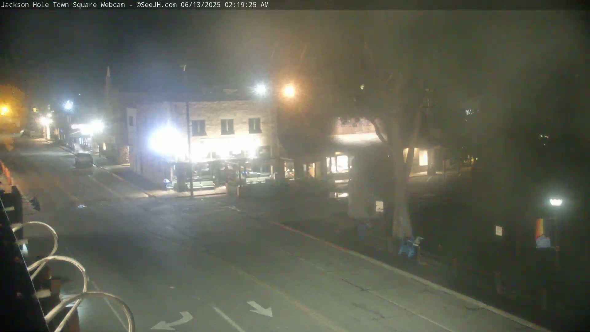 A nighttime view of a mostly empty town square with a few vehicles parked and some buildings visible under hazy conditions.