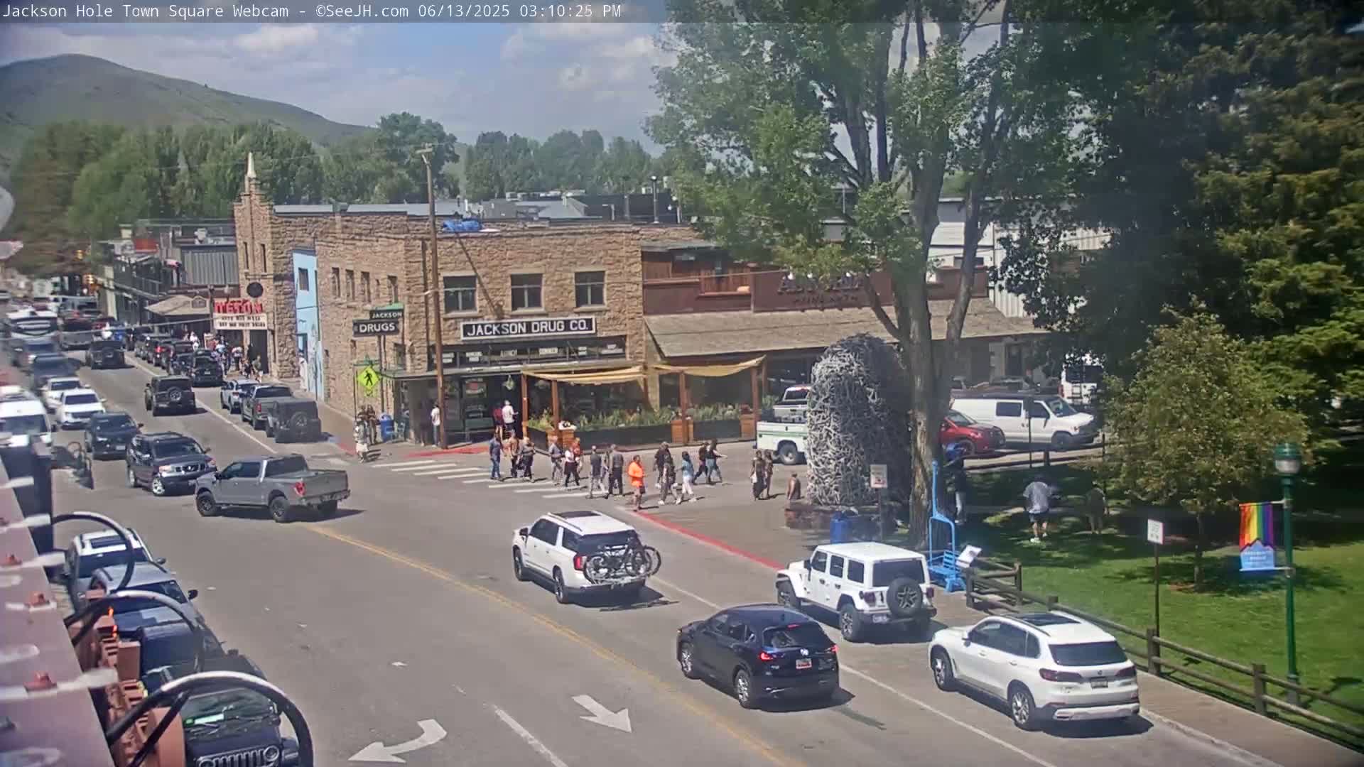 A sunny day in a town square with numerous cars parked and driving on the street, pedestrians crossing a crosswalk in front of several buildings, and a grassy area with trees in the background.