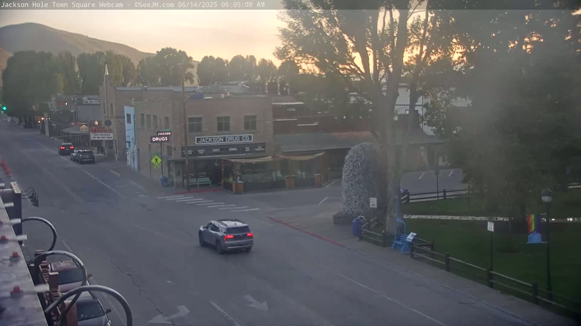 A hazy sunrise illuminates a town square with several buildings, including one labeled "Jackson Drug Co," and a few cars driving on the road.
