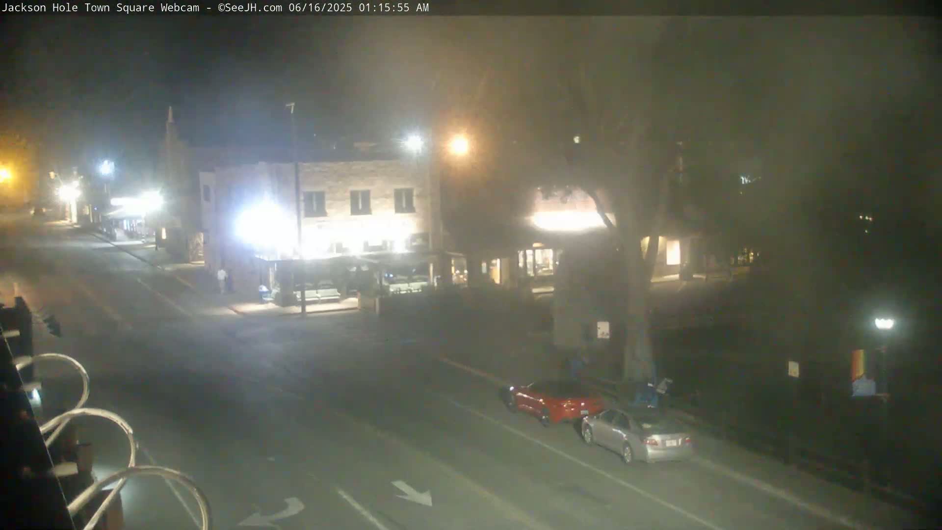 A nighttime view of a town square with two cars parked on the street, several buildings with lights on, and a hazy atmosphere.