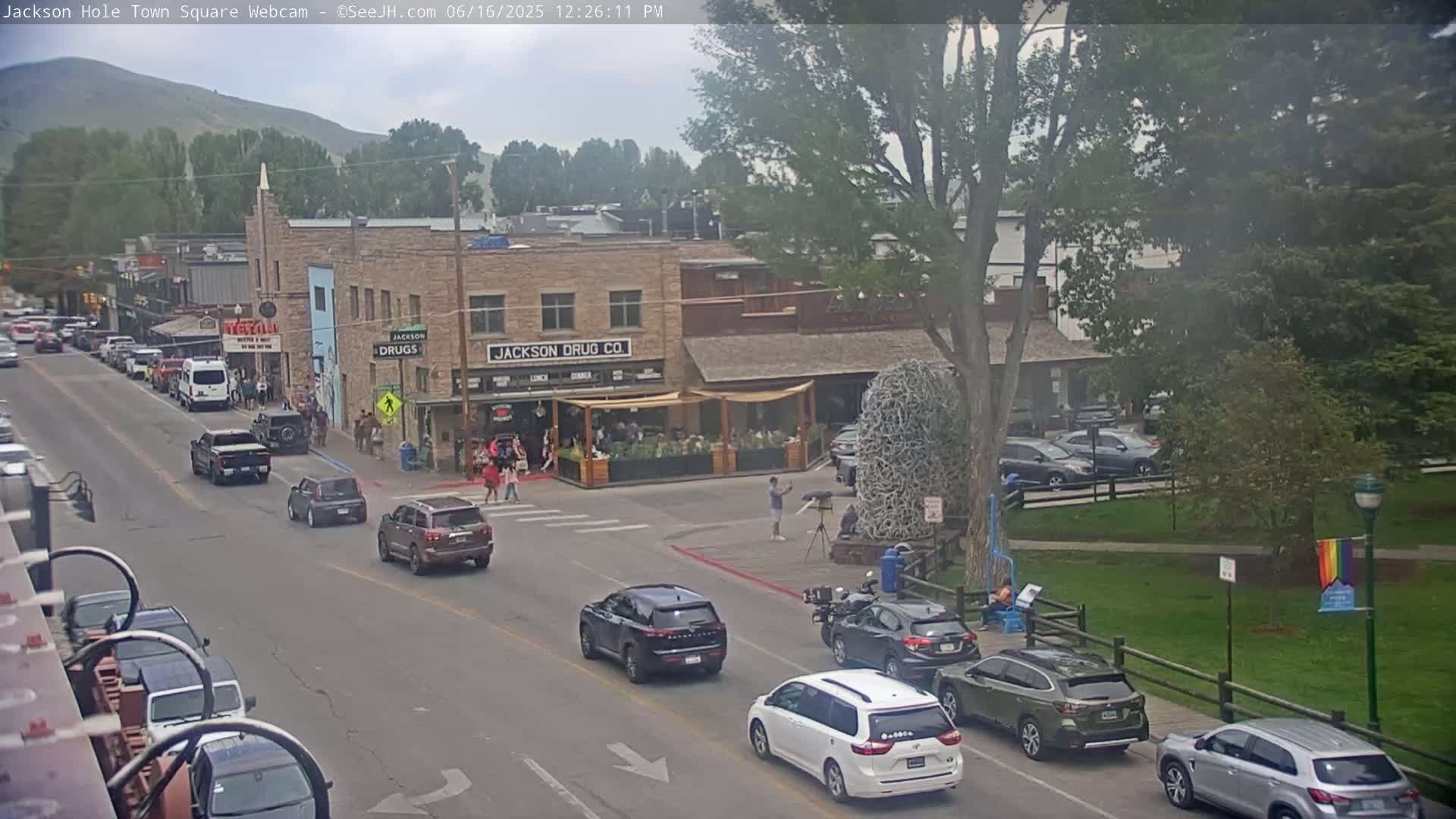 A street scene in a town shows several cars driving past buildings with pedestrians on the sidewalk, under an overcast sky.