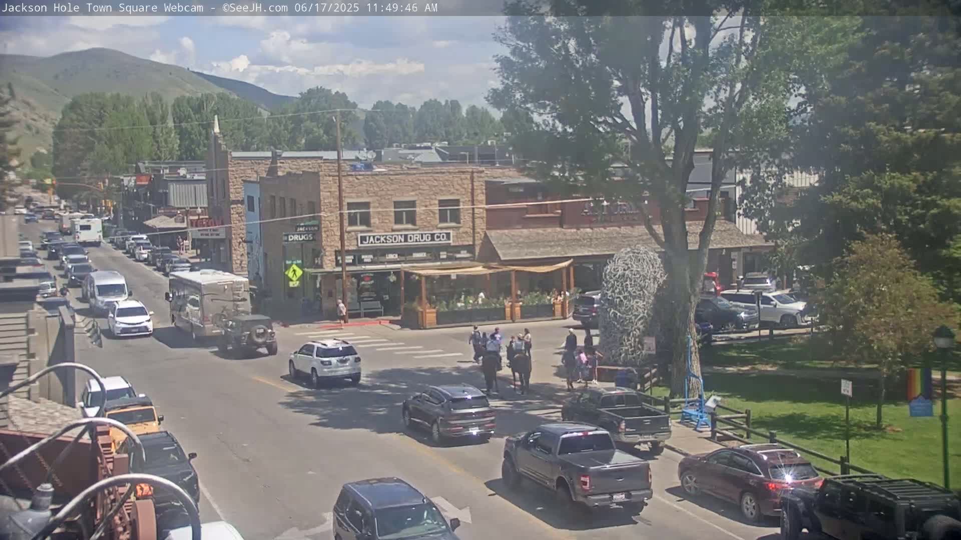 A sunny day in a town square shows several cars driving on a street lined with buildings, some pedestrians and a horse-mounted individual crossing the street near a grassy area.