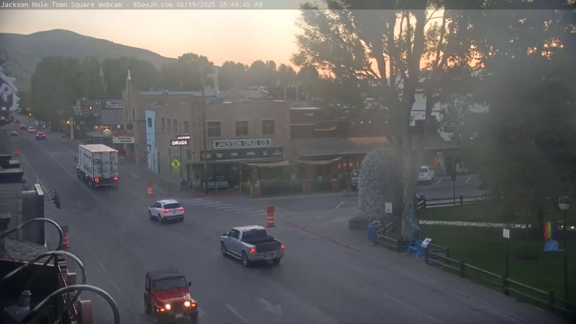 A hazy sunrise illuminates a town square with several vehicles on the road, including a large truck, SUV, pickup truck, and jeep.