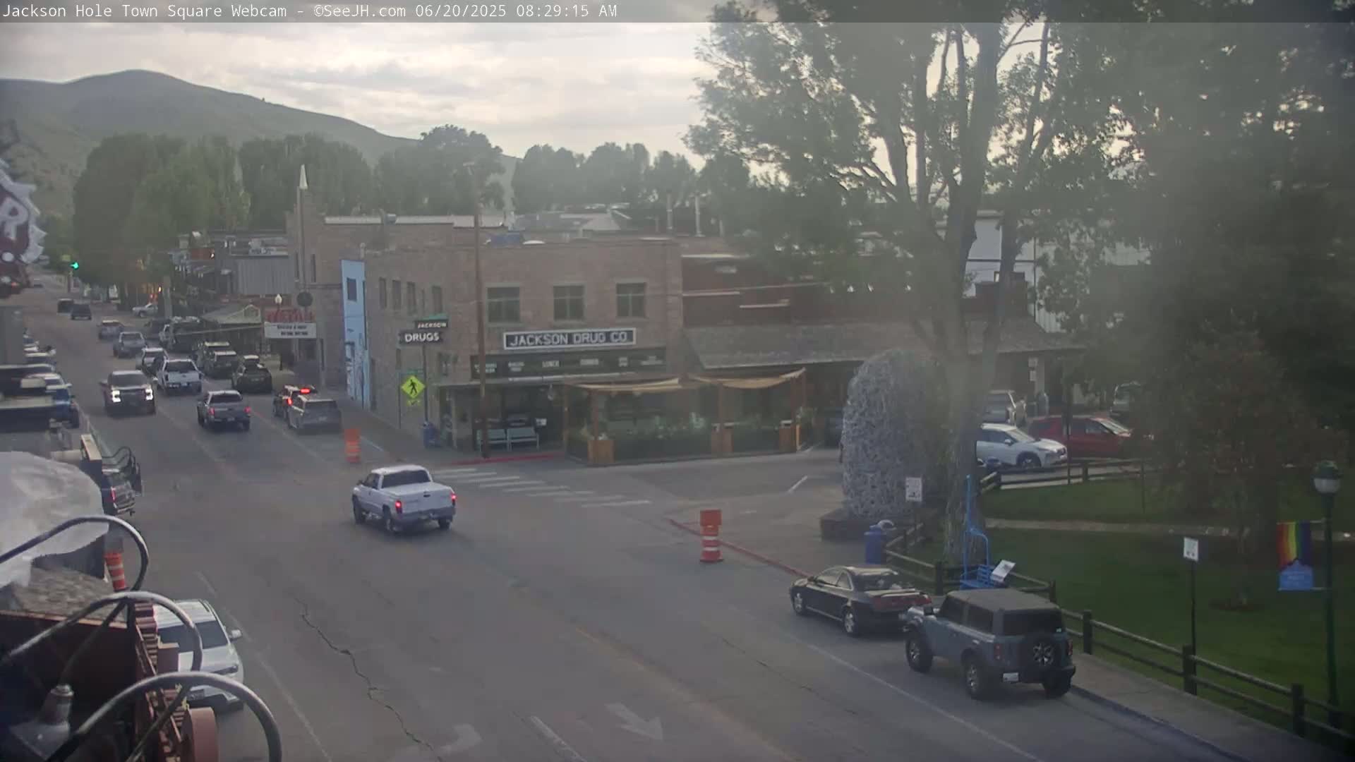 A hazy morning view of a town square, showing several vehicles on a street in front of various buildings, including one labeled "Jackson Drug Co," with a park area visible to the right.