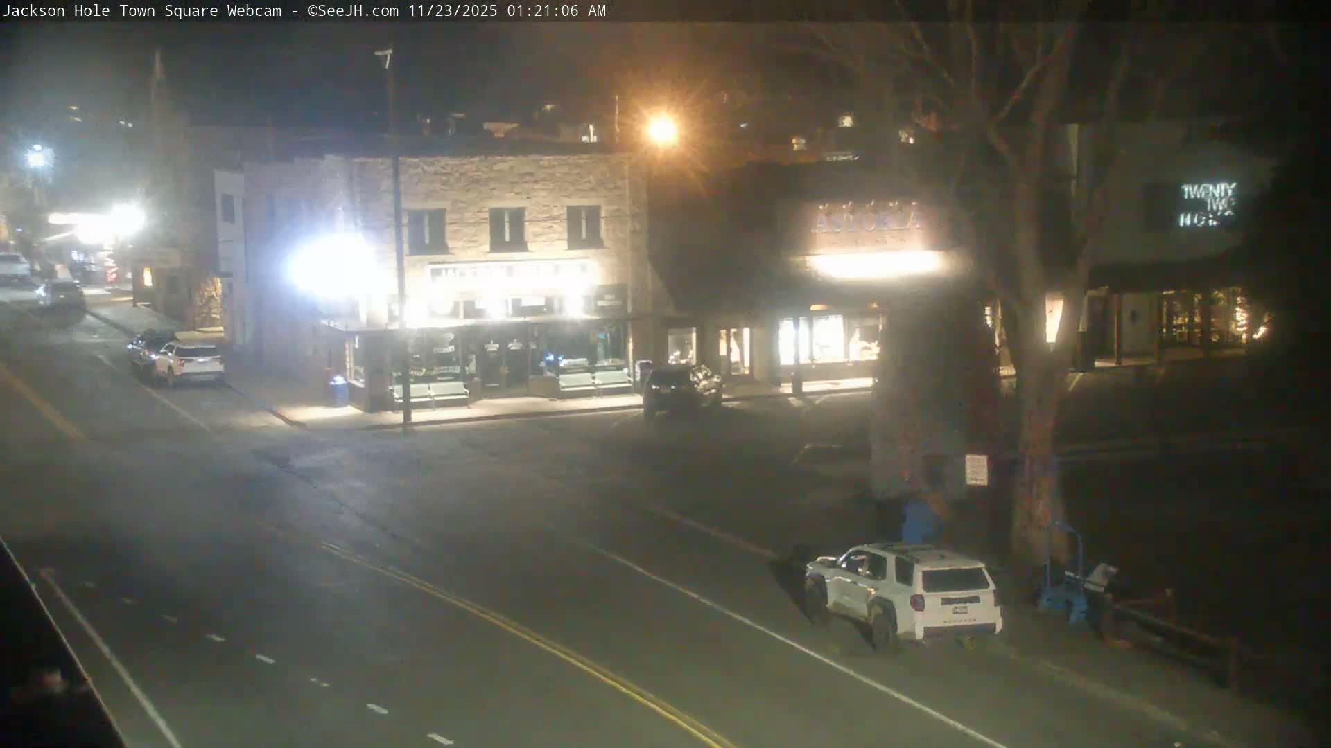 A clear and dry nighttime view captures a brightly lit town street flanked by multi-story stone buildings with illuminated storefronts, several cars parked and driving, and a large bare tree on the right.