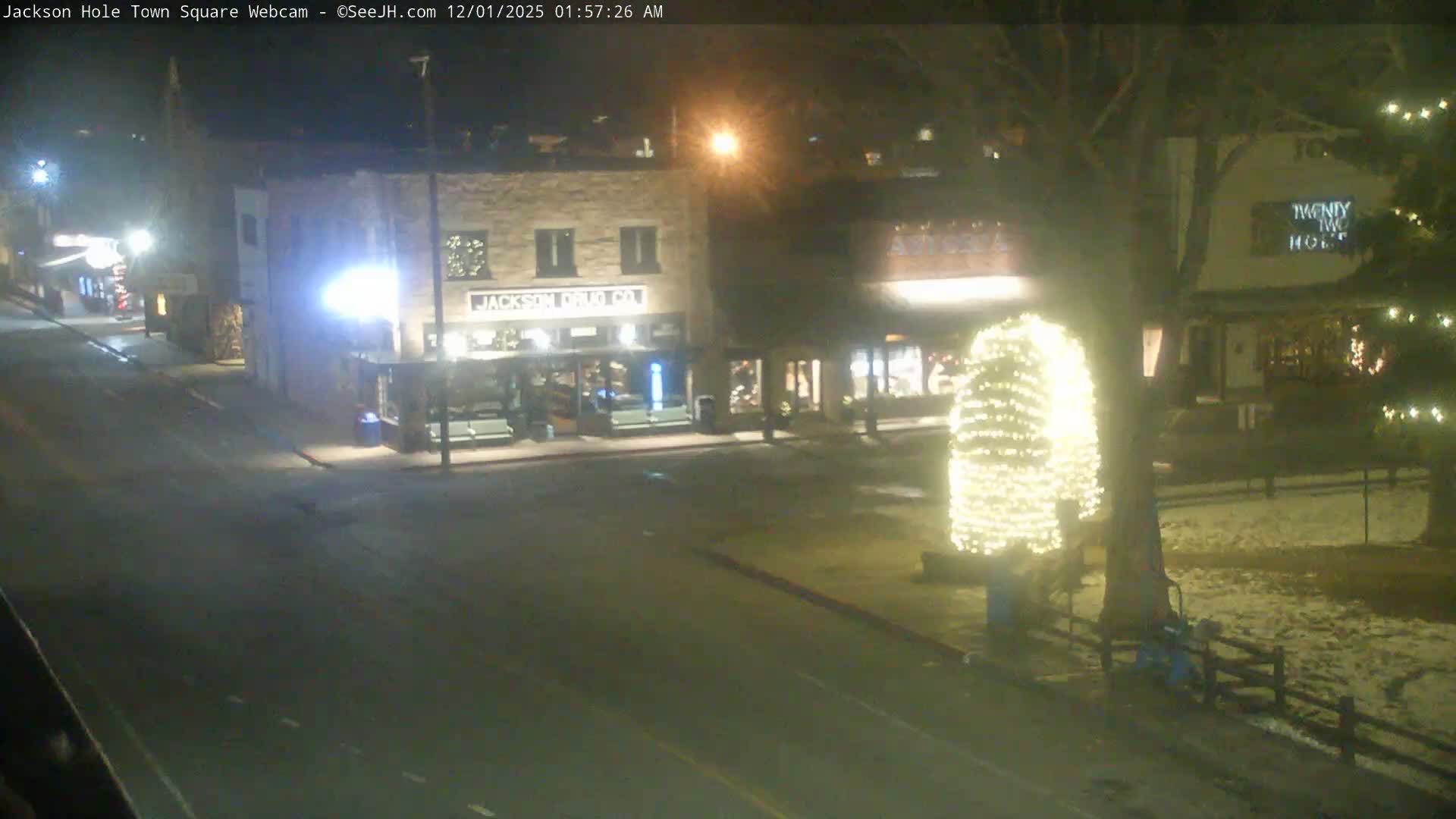 A quiet town square is brightly lit at night with buildings and a large tree adorned with warm white string lights, while the ground is partially covered in snow, indicating cold, clear weather.