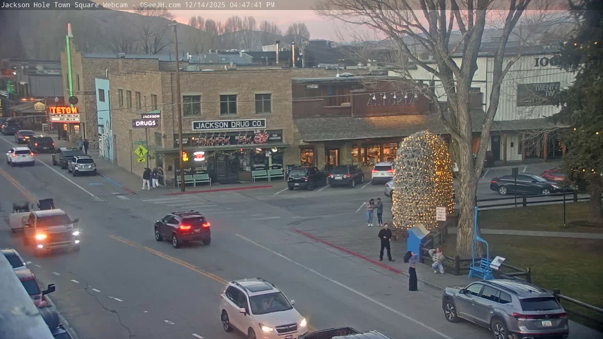 A quiet, snow-covered town square at night is illuminated by bright streetlights and storefront windows, revealing buildings and a prominent bare tree under clear, cold conditions.