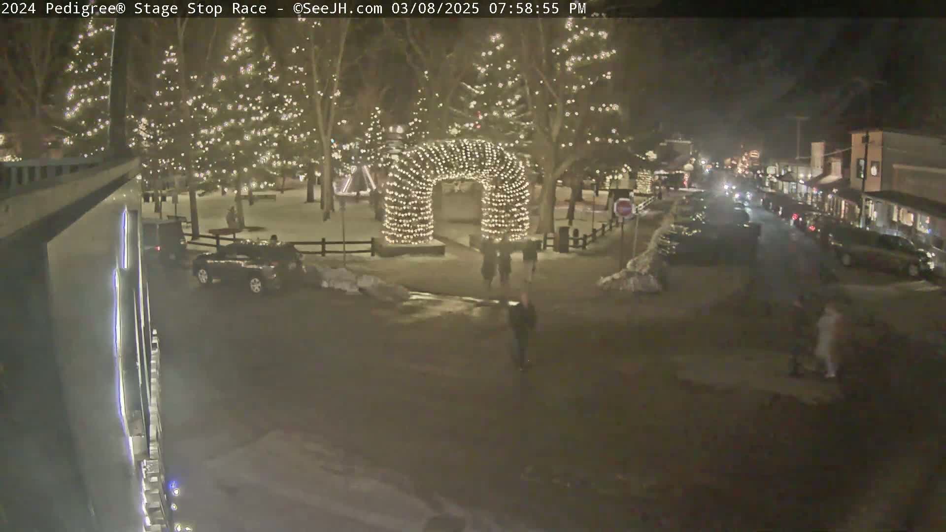 A nighttime scene shows a snow-covered town square with a lighted archway, several cars parked along the streets, and people walking around under the glow of holiday lights on trees.