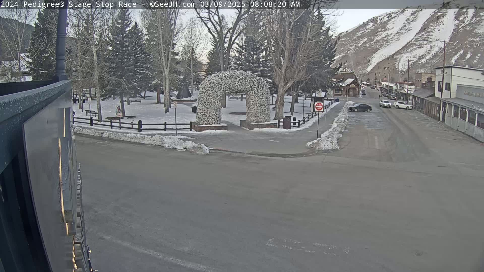 A snow-covered town square with a large antler archway is visible, and a street with several parked cars leads to buildings up a snowy mountainside.