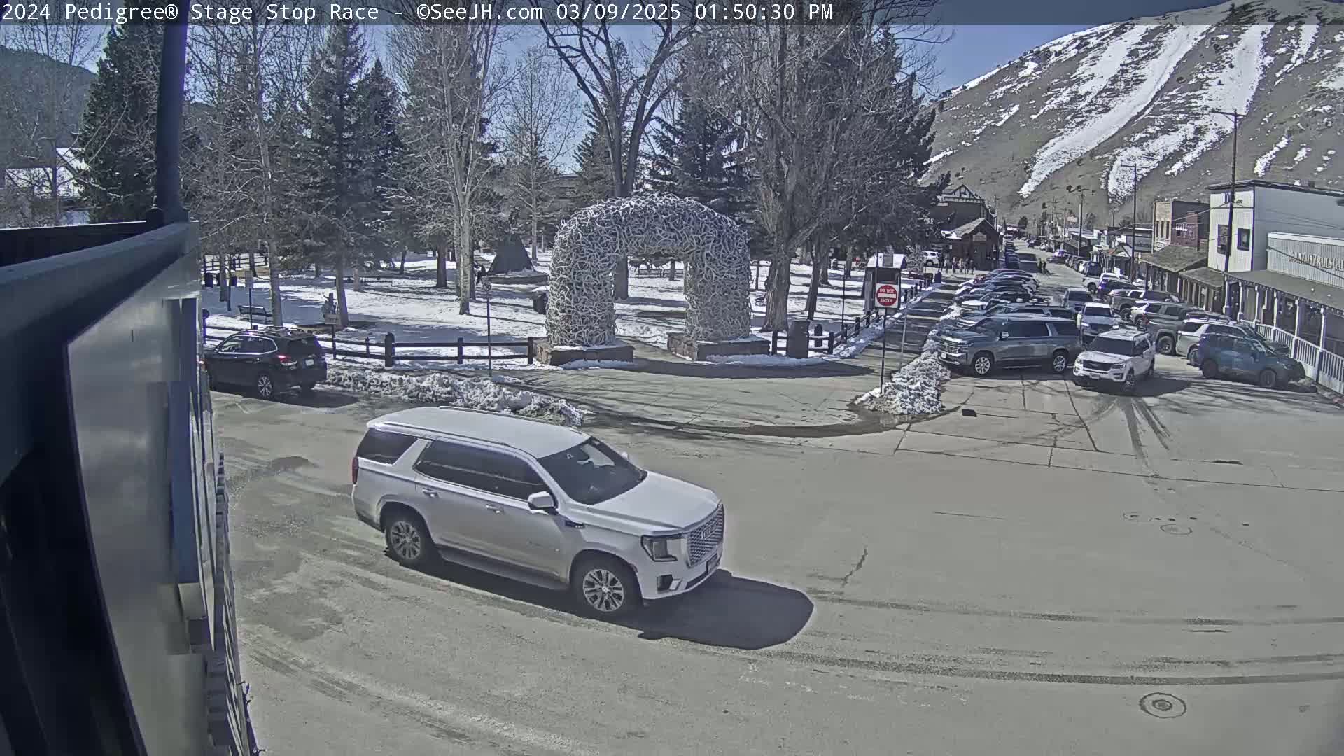 A sunny day reveals a snow-dusted town square with parked cars, a large antler archway, and a silver SUV driving by.