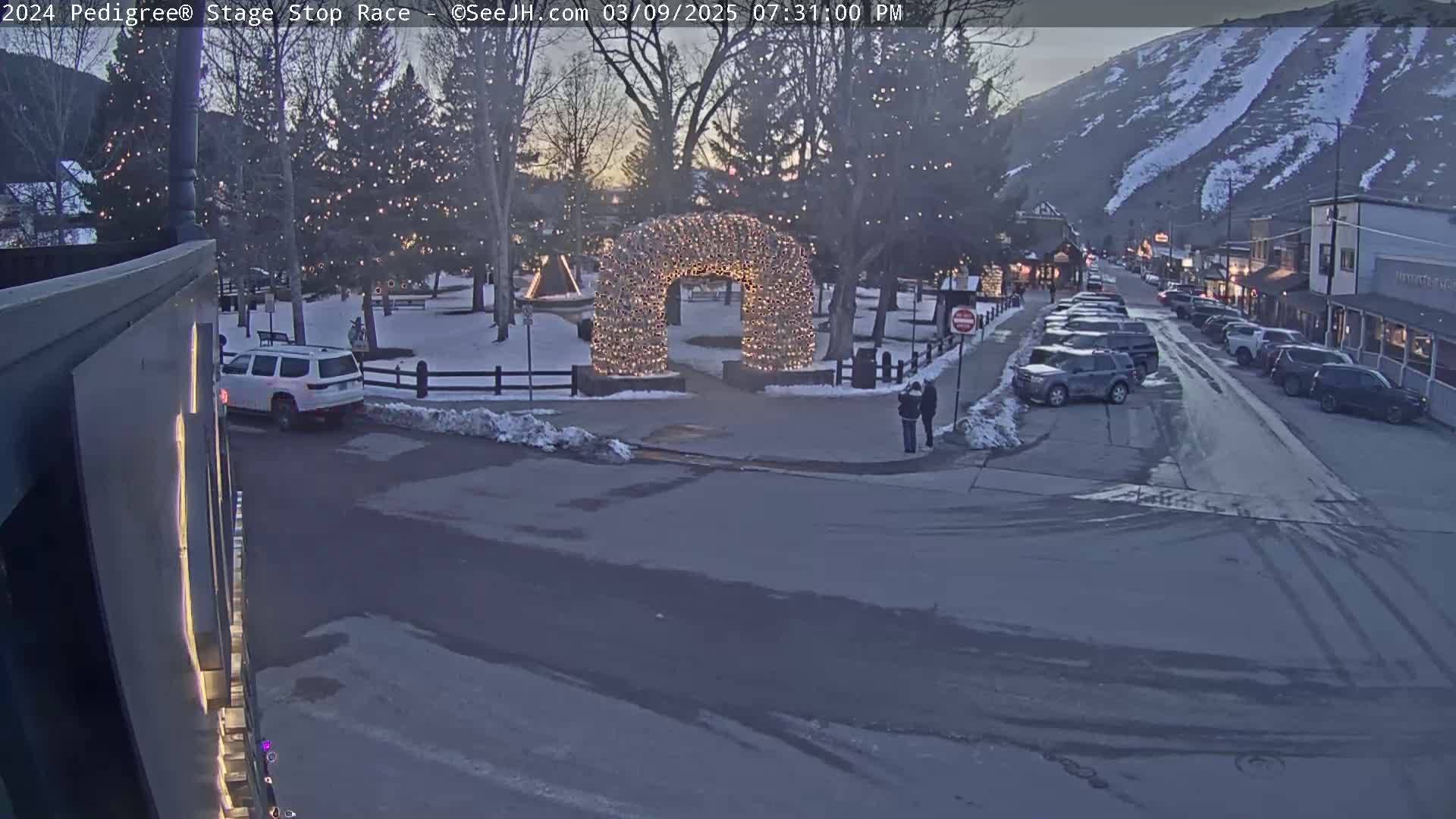 A snow-dusted town square at dusk features a light-covered archway, parked cars lining a street, and a mountain in the background.