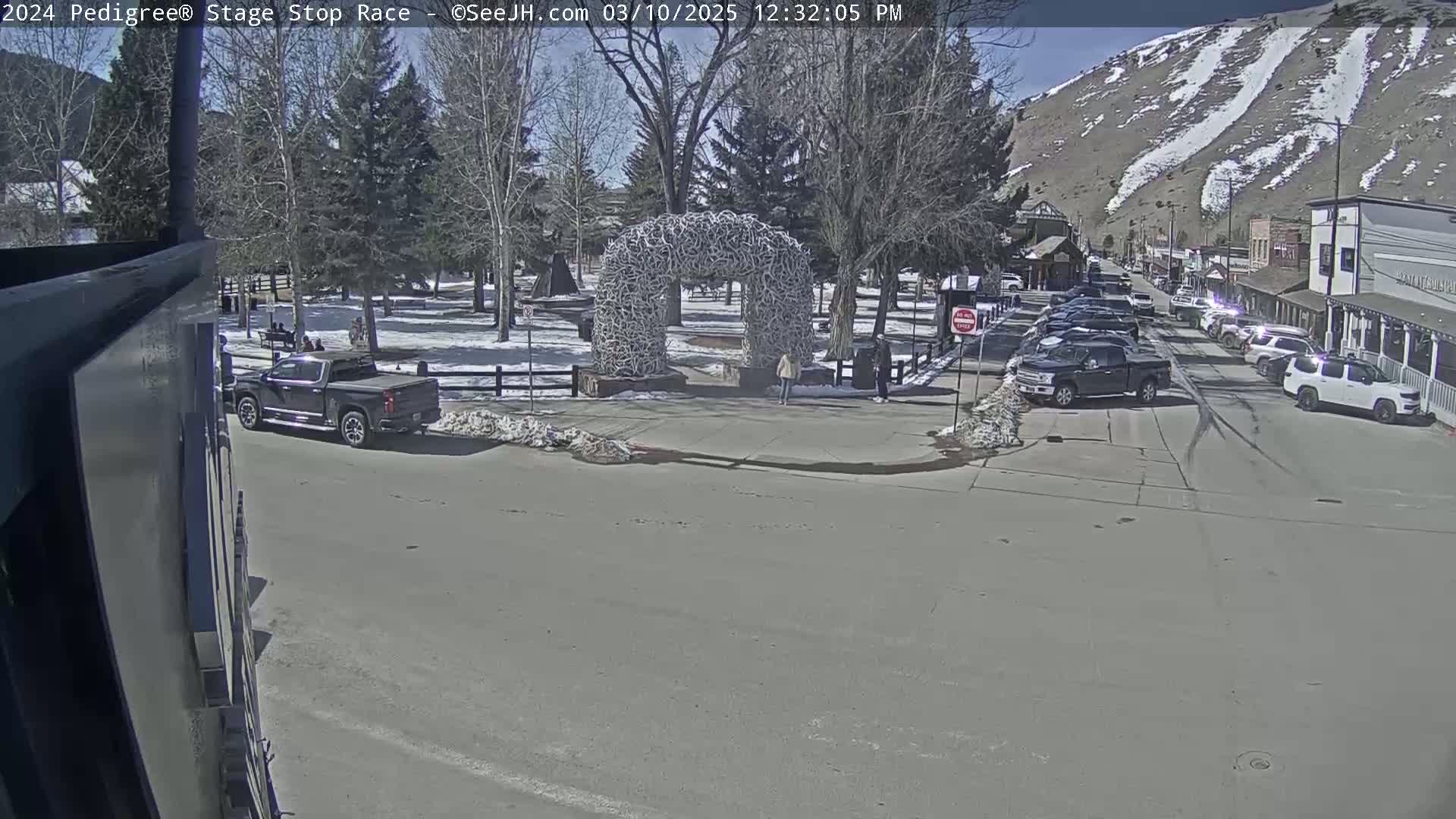 A sunny day shows a street scene with parked cars, a pickup truck, and a large antler arch in a snow-dusted park, all set against a backdrop of mountains.