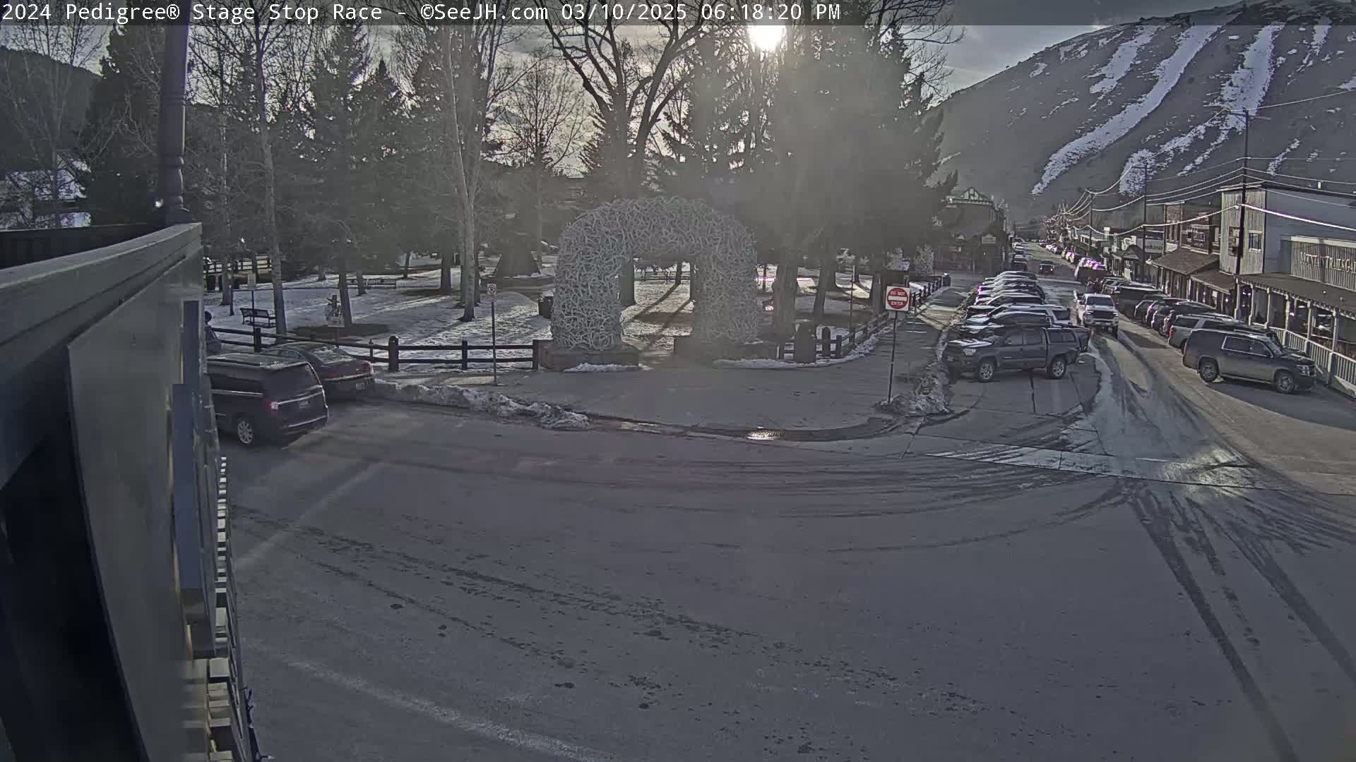 A sunny, winter day reveals a snow-dusted town square with parked cars lining a street beside a mountain, featuring an archway constructed of antlers in the center.