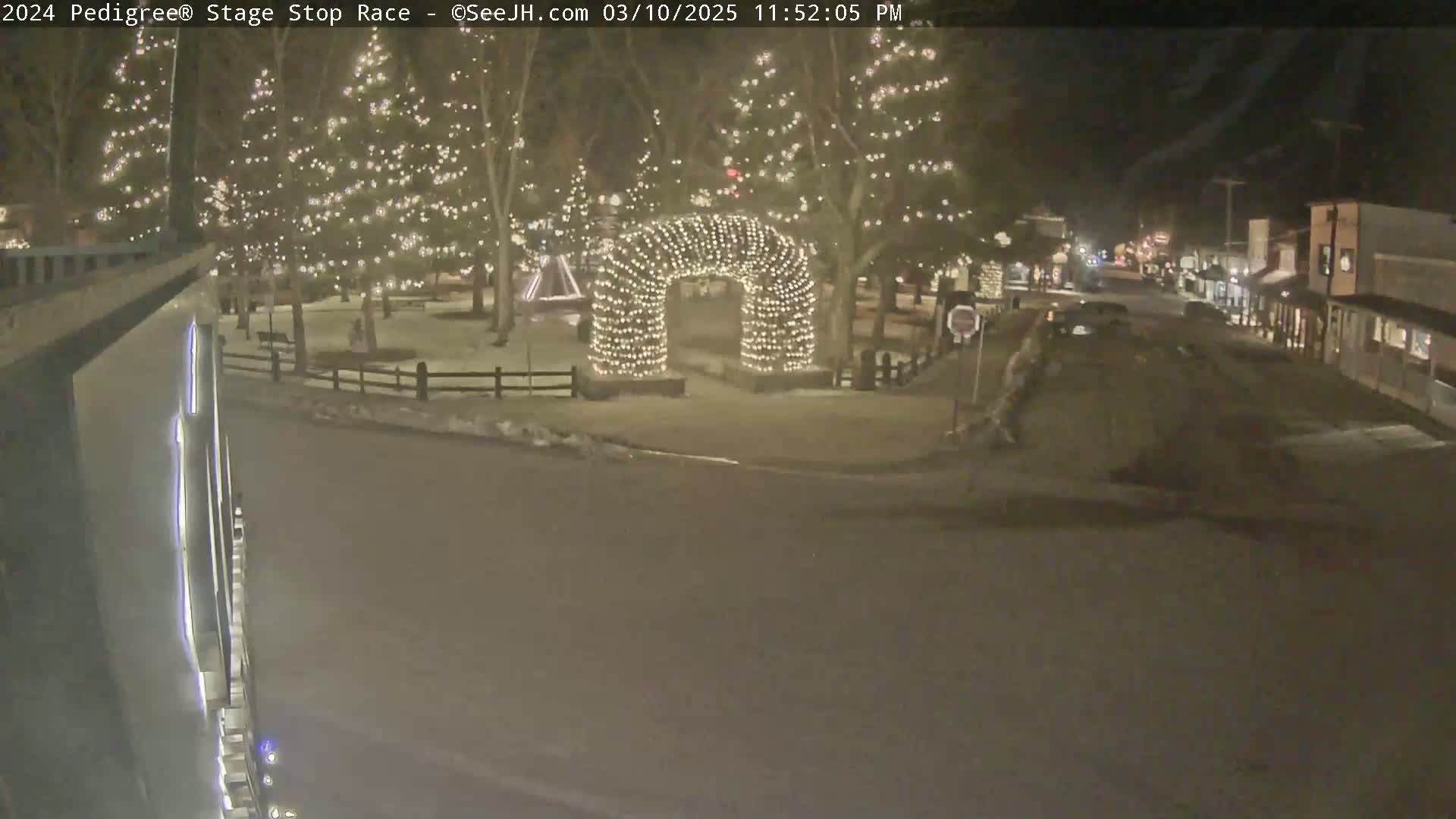 A nighttime scene shows a snow-covered town square with Christmas lights illuminating trees and an archway, with a street and buildings visible in the background.