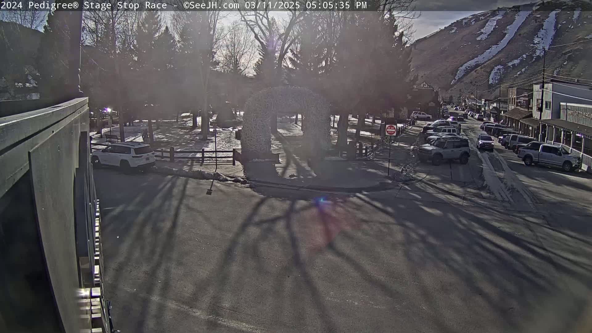 A sunny winter day shows a town square with a decorative arch, parked cars lining a street, and a mountainside with a ski run in the background.