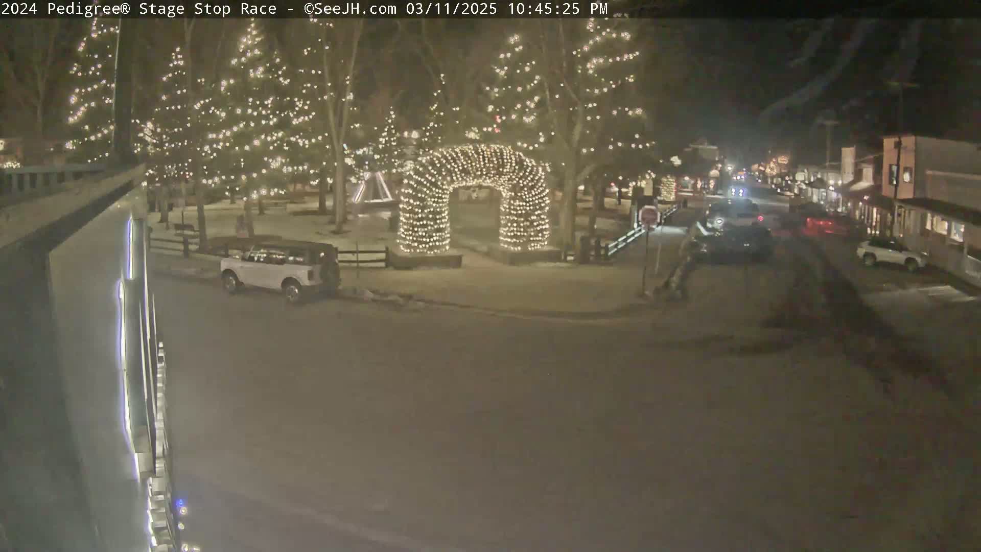 A nighttime snowy scene shows a town square with illuminated trees and an archway of lights, along with cars parked and driving on a street lined with buildings.