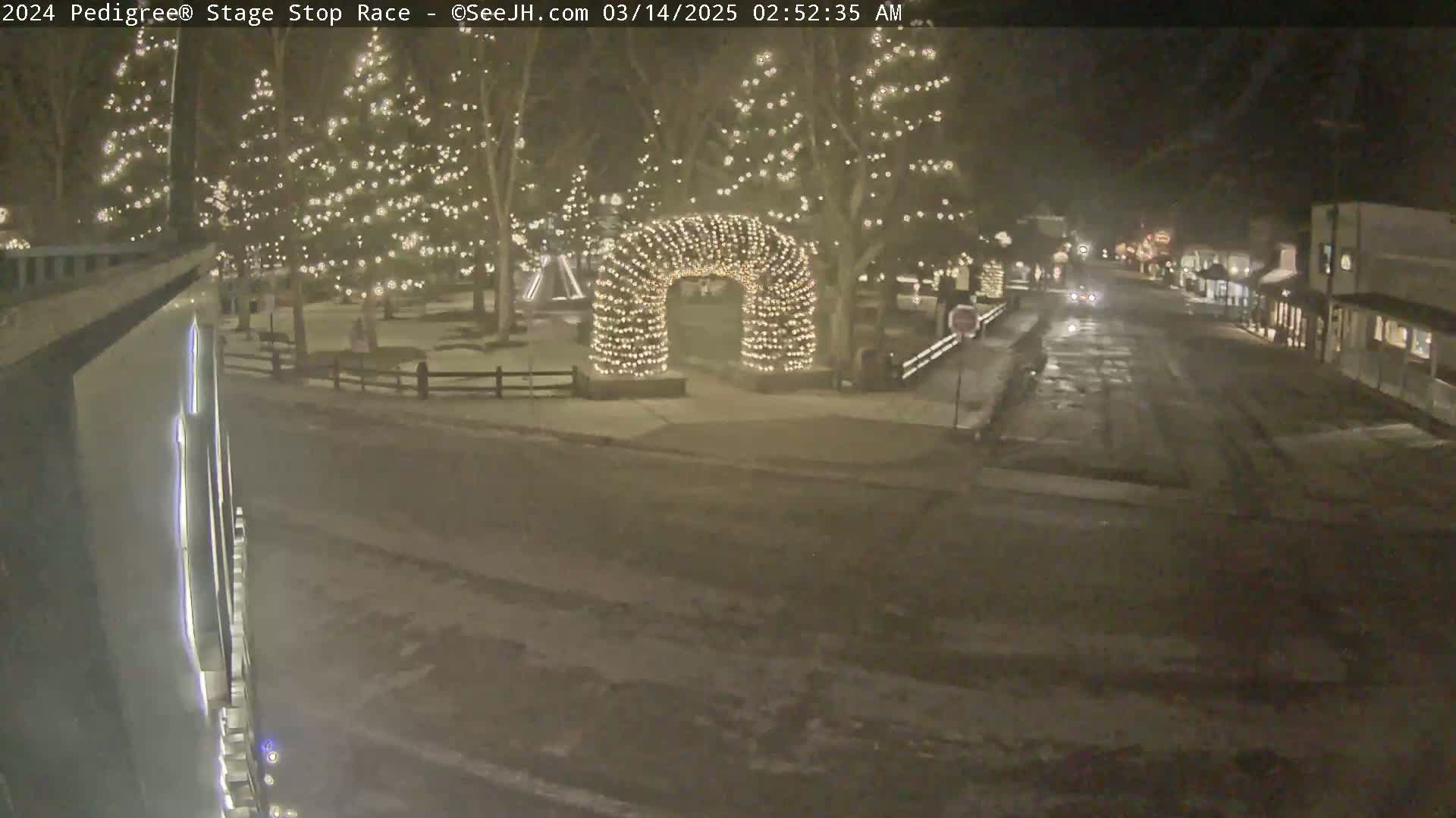 A snow-covered street at night, illuminated by Christmas lights strung on trees and forming an archway, with a few vehicles visible in the distance.