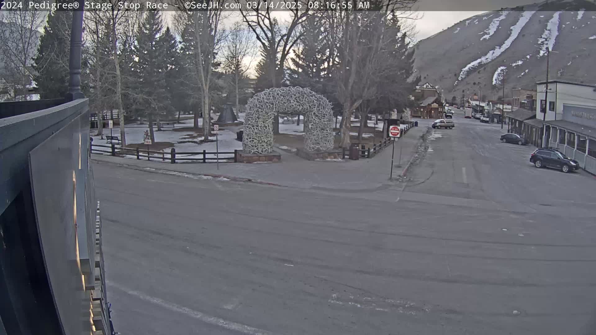 A mostly empty street in a town with snow-covered ground and mountains in the background, featuring a large antler archway in a park-like setting.