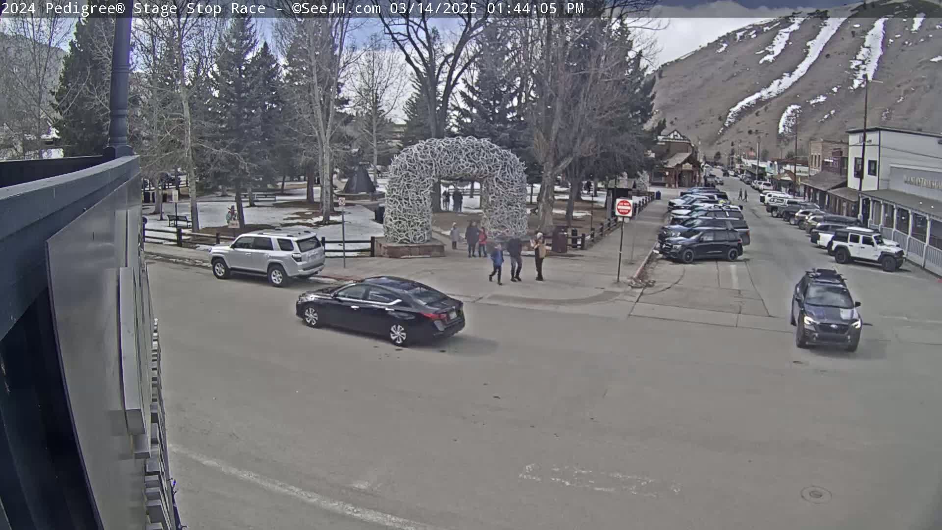 A town square with several cars parked along the street, a large antler archway in the center, and a few people walking around on a partly cloudy day with snow-covered mountains in the background.