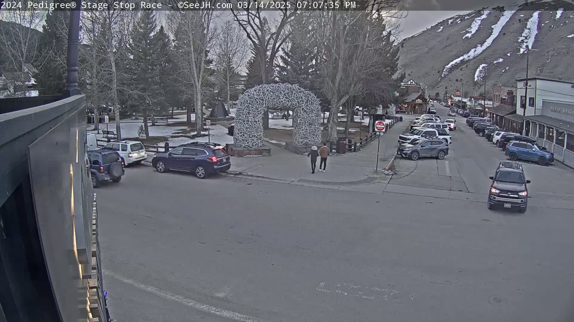 A street scene shows several cars parked along the sides of a street, with a few people walking near a large antler arch in a snowy park, all under an overcast sky.