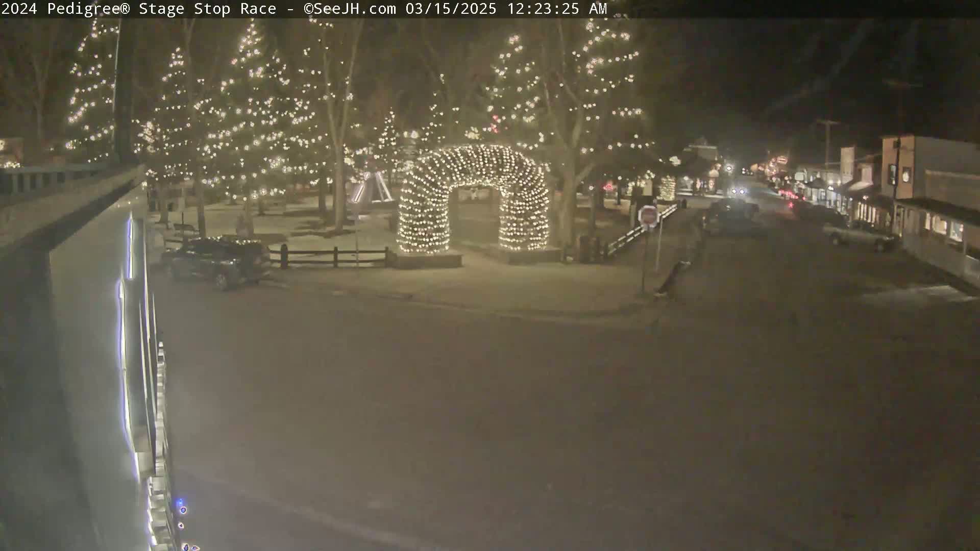 A nighttime scene shows a snow-covered town square with illuminated trees and a light-covered archway, and a street with cars driving by.