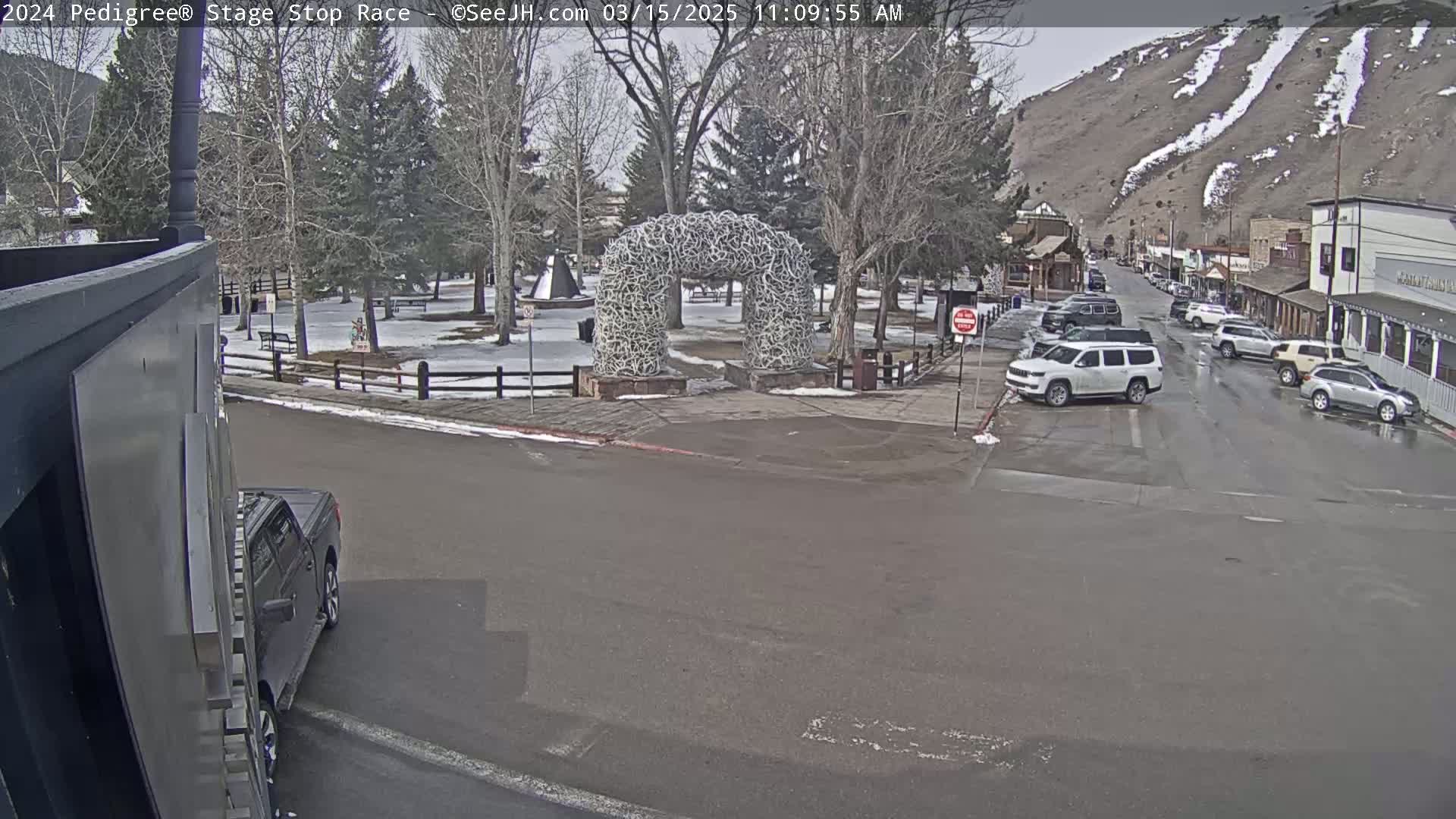 A town square with a large antler archway is visible, with several parked cars and a few driving down the street on an overcast day.
