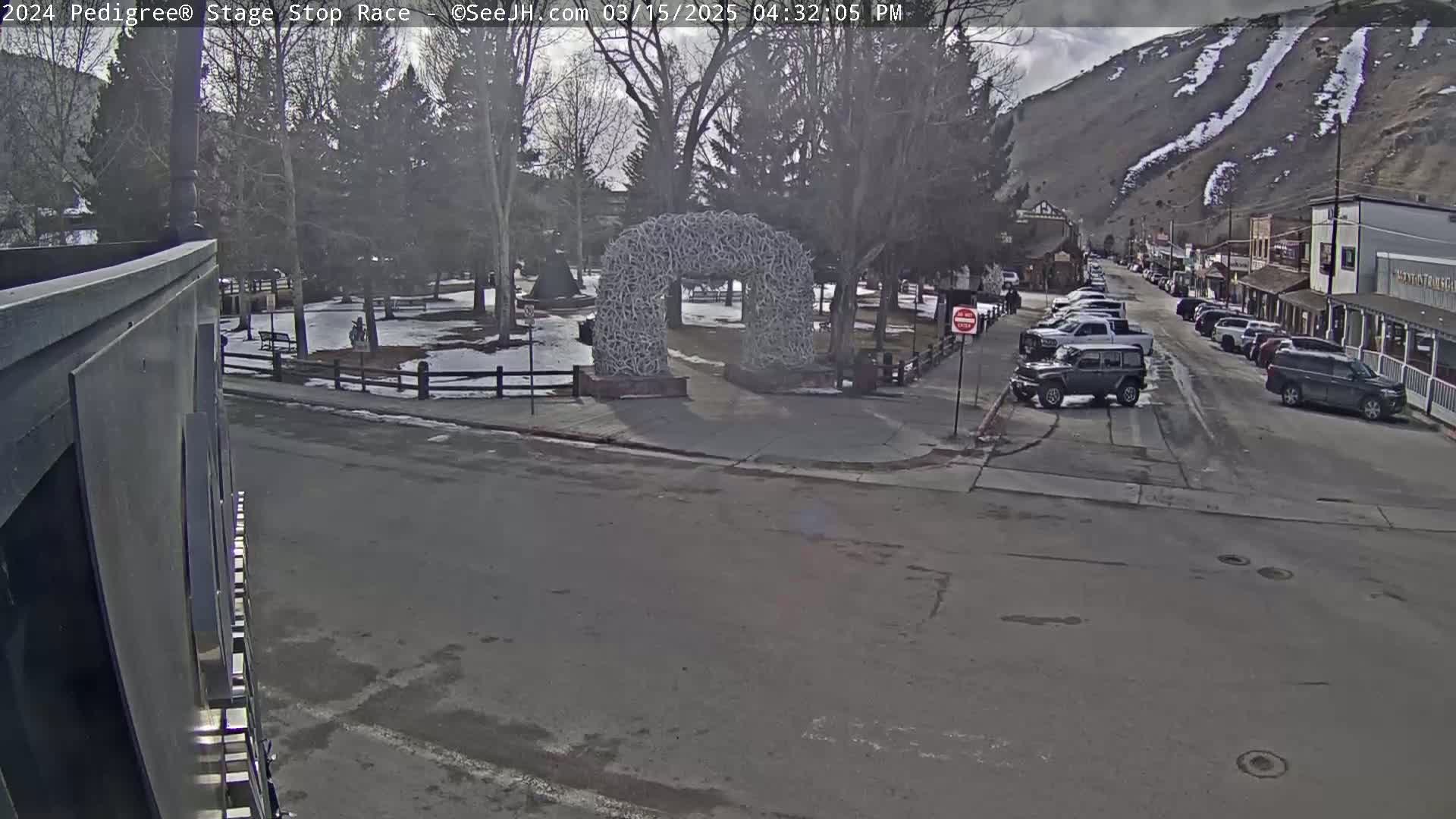 A street scene on a partly cloudy day shows a town square with a large antler archway, parked cars lining a street that leads up to a mountainside with snow streaks.