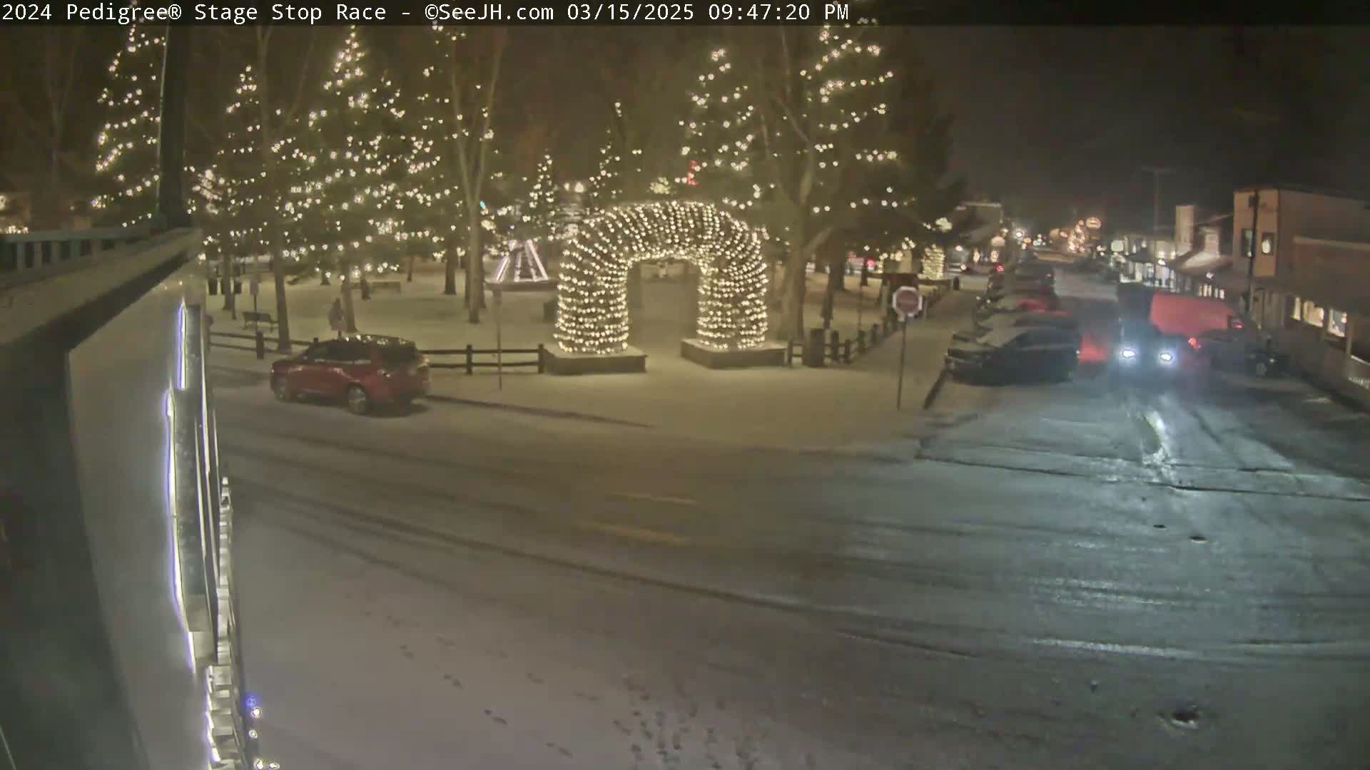 A snow-covered town square at night, illuminated by Christmas lights strung on trees and an archway, with several cars parked and driving on nearby streets.