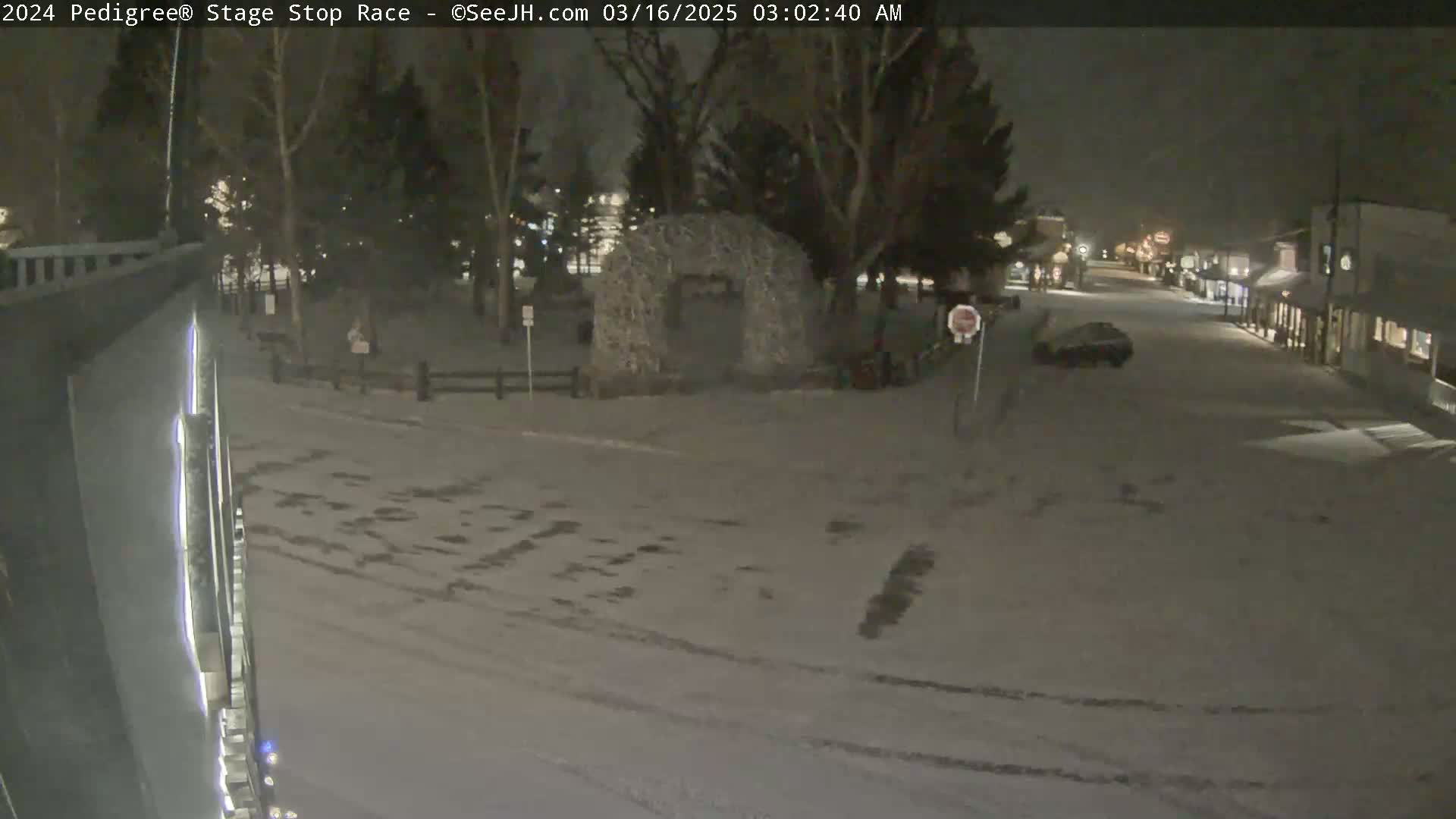 A snow-covered town square at night, with a stone archway and a few parked cars visible under dim lighting.