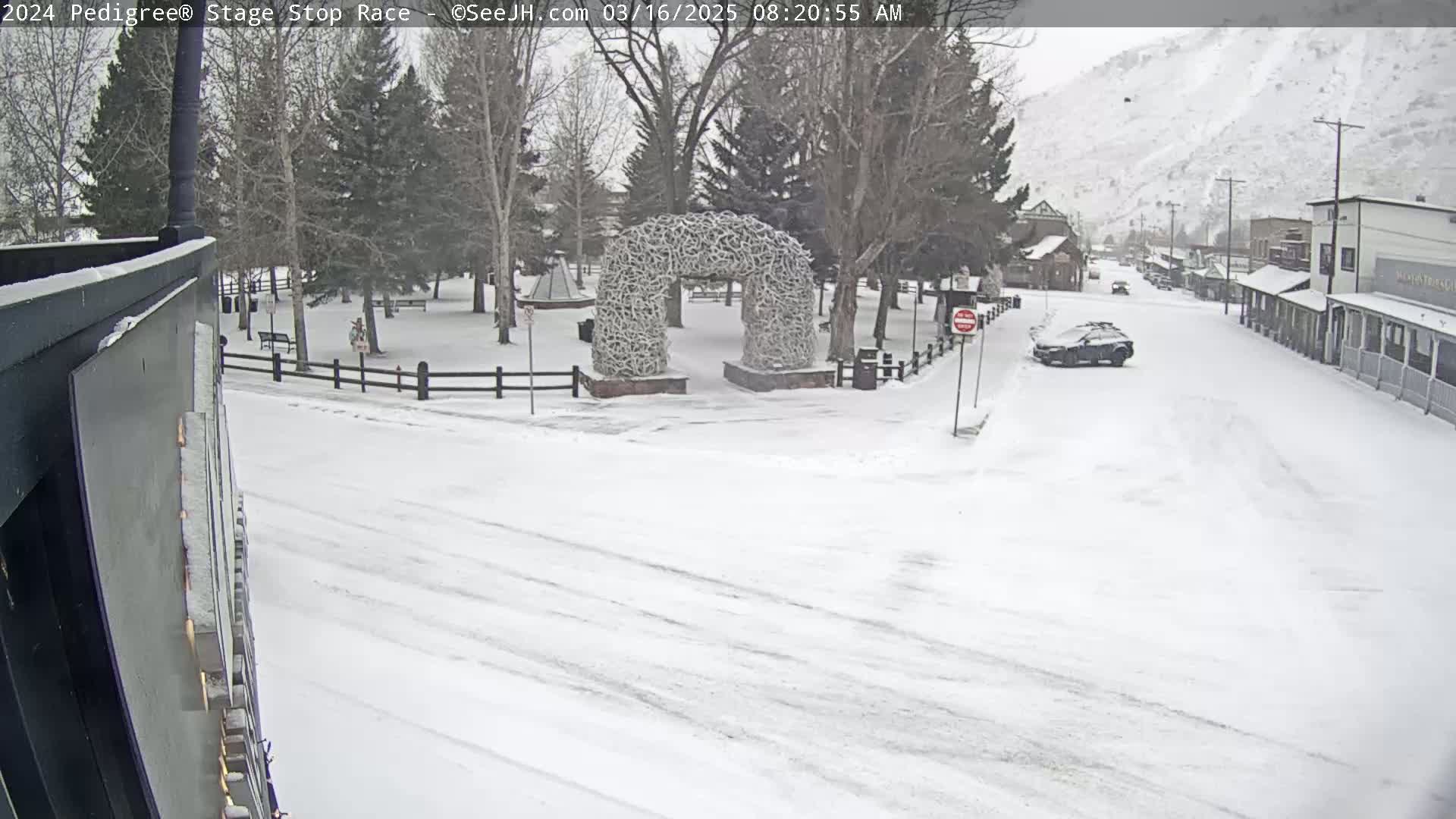A snow-covered town square features a large antler archway, a parked car, and a few buildings in the background under a snowy sky.