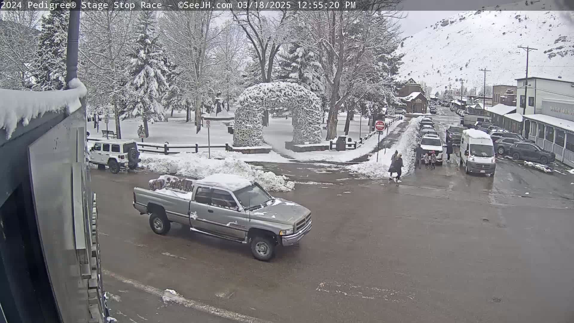 A snow-covered town square with several vehicles, including a pickup truck and a van, and pedestrians crossing a street.