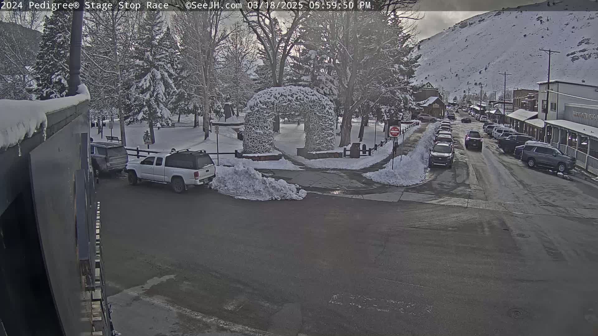 A snow-covered town square with parked cars and a snow-covered archway, under an overcast sky.