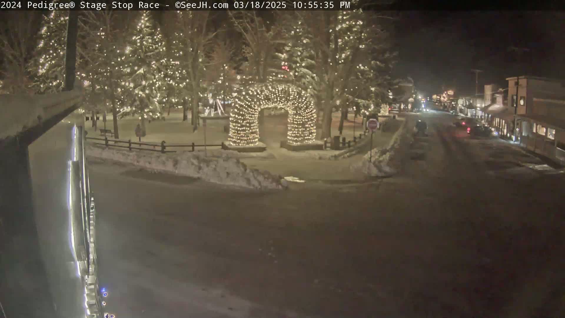 A snow-covered town square at night, illuminated by string lights strung in an archway and on trees lining the square, with a street visible beyond.