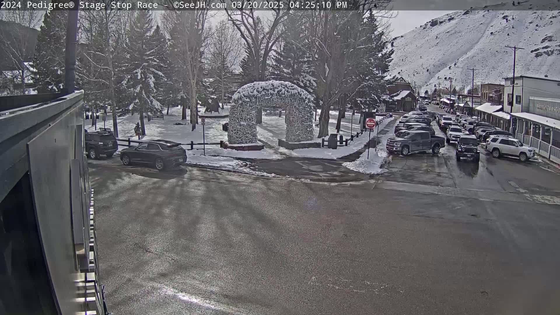 A snow-covered town square with a stone archway, several cars parked along the street, and a background of snow-dusted mountains.