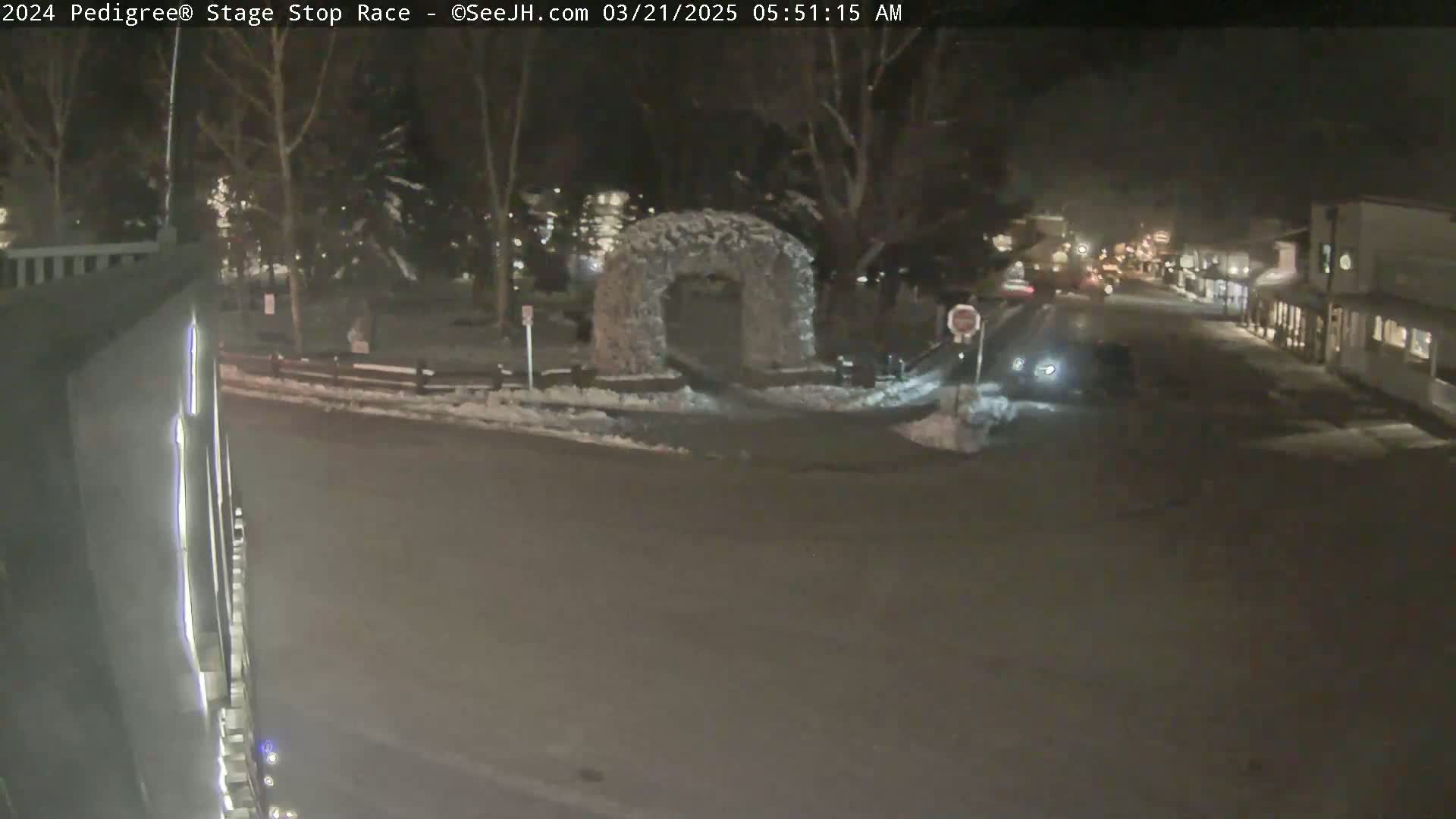 A snow-covered street scene at night shows a few cars, buildings, and a snow-covered archway in a park-like setting.