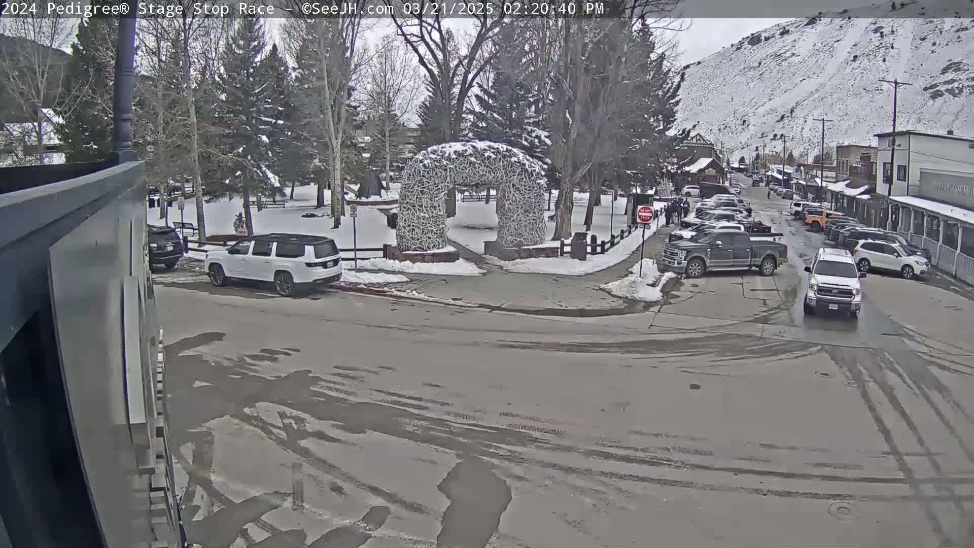 A snowy town square with several vehicles parked and driving around a snow-covered archway, under an overcast sky.