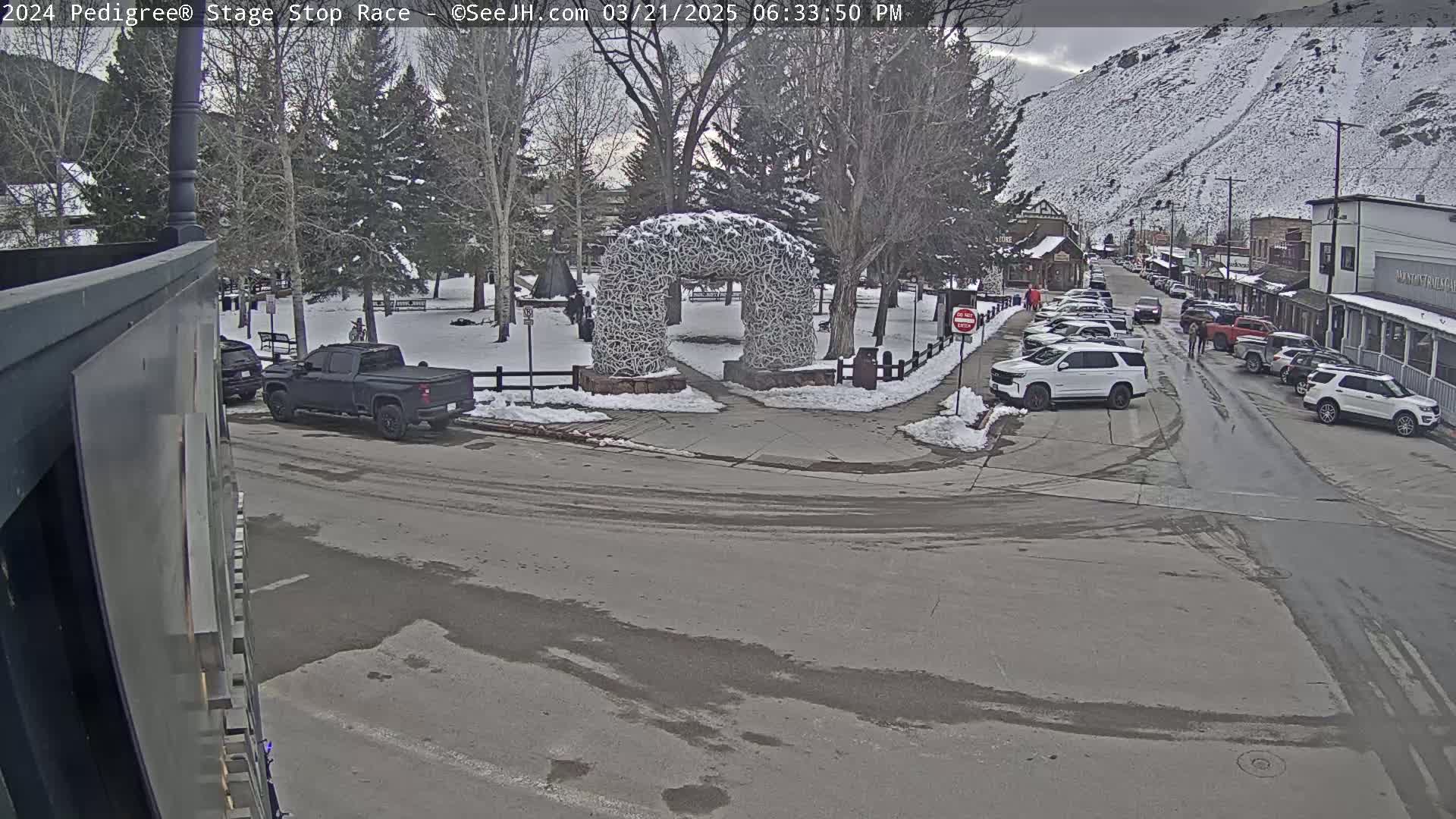 A snowy mountain town scene shows a street with parked cars, a park with an arch made of antlers, and a few pedestrians.