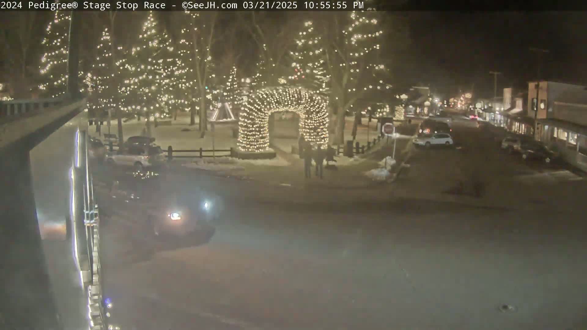 A nighttime, snow-covered town square features Christmas lights strung on trees and an archway, with several cars parked on a nearby street.