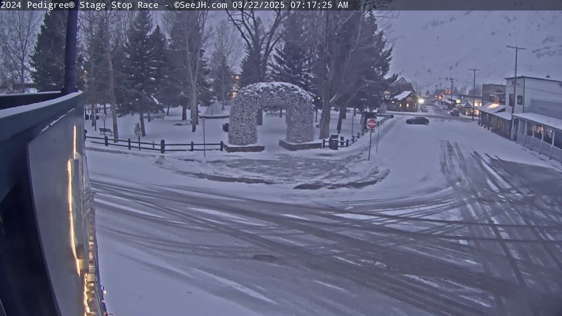A snow-covered town square with a stone archway features tire tracks on the roads, and a few vehicles are visible in the distance under a cloudy sky.
