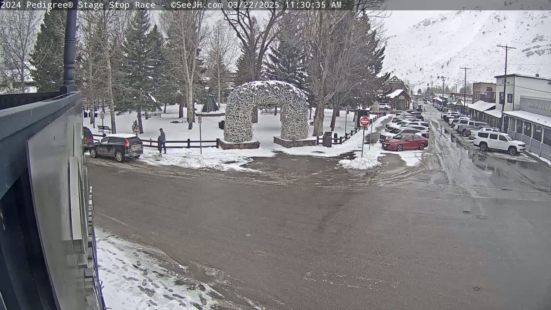 A snow-covered town square with a large antler archway, several parked cars, and a few pedestrians, under a cloudy sky.