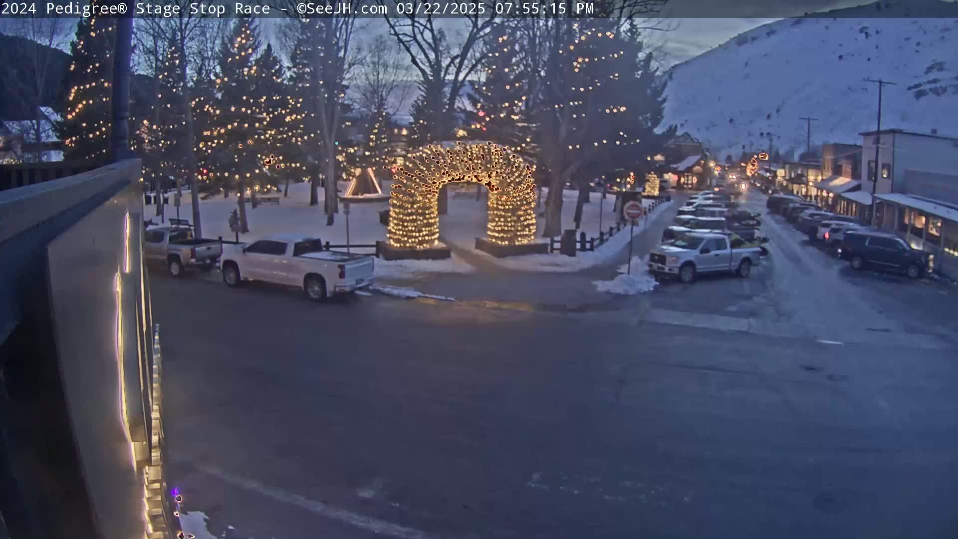 A town square at dusk, illuminated by Christmas lights on trees and an archway, with parked cars and snow on the ground.