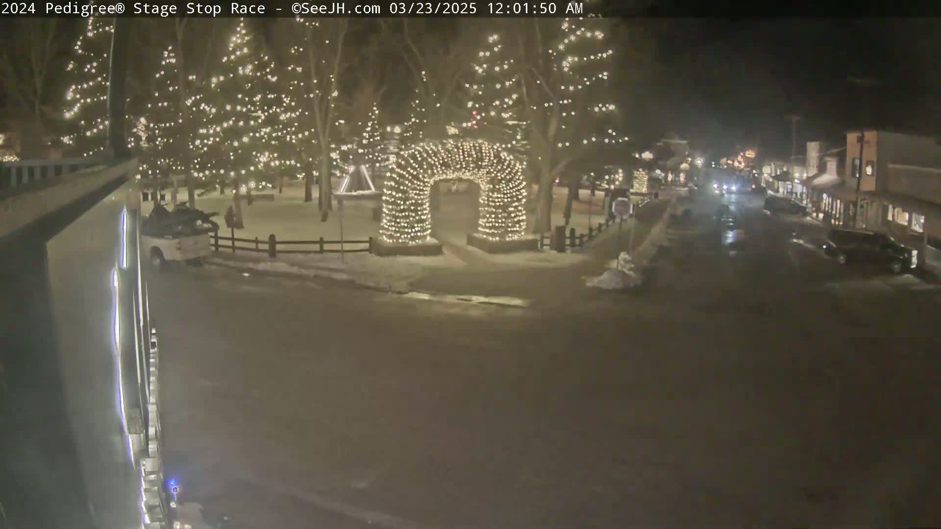 A nighttime scene shows a snow-covered town square with Christmas lights illuminating trees and an archway, while a few vehicles are visible on a nearby street.