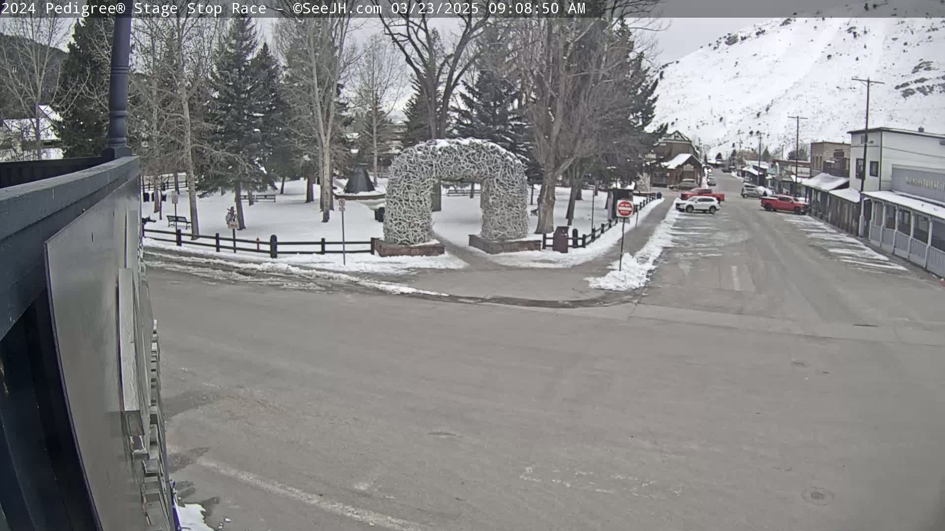 A snow-covered town square with a unique archway made of antlers is visible from a street view, with cars parked along the sides of a road leading to buildings nestled against a snow-dusted mountain.